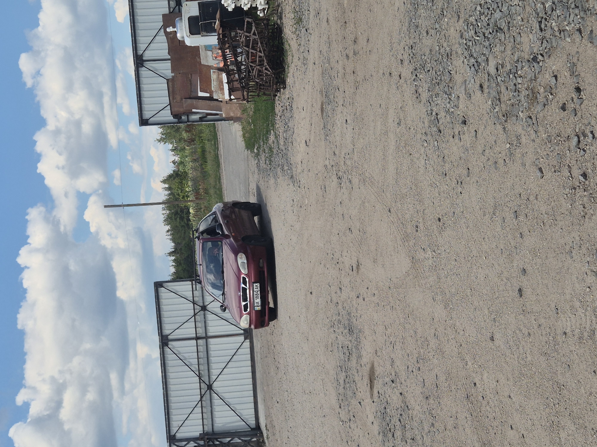 A burgundy Daewoo Lanos sedan with license plate BA 1884 AK is parked on a gravel lot. The car is facing right and slightly angled towards the viewer. Behind the car, a large white corrugated metal gate is visible on the left side of the image. To the right of the car, there is another building or structure with corrugated metal siding, and a pile of rusty metal farm equipment is stacked in front of it. The environment appears to be an outdoor industrial or agricultural area, possibly a junkyard or storage facility, under a partly cloudy sky. The location is identified as Pidhaitsi, Ukraine.