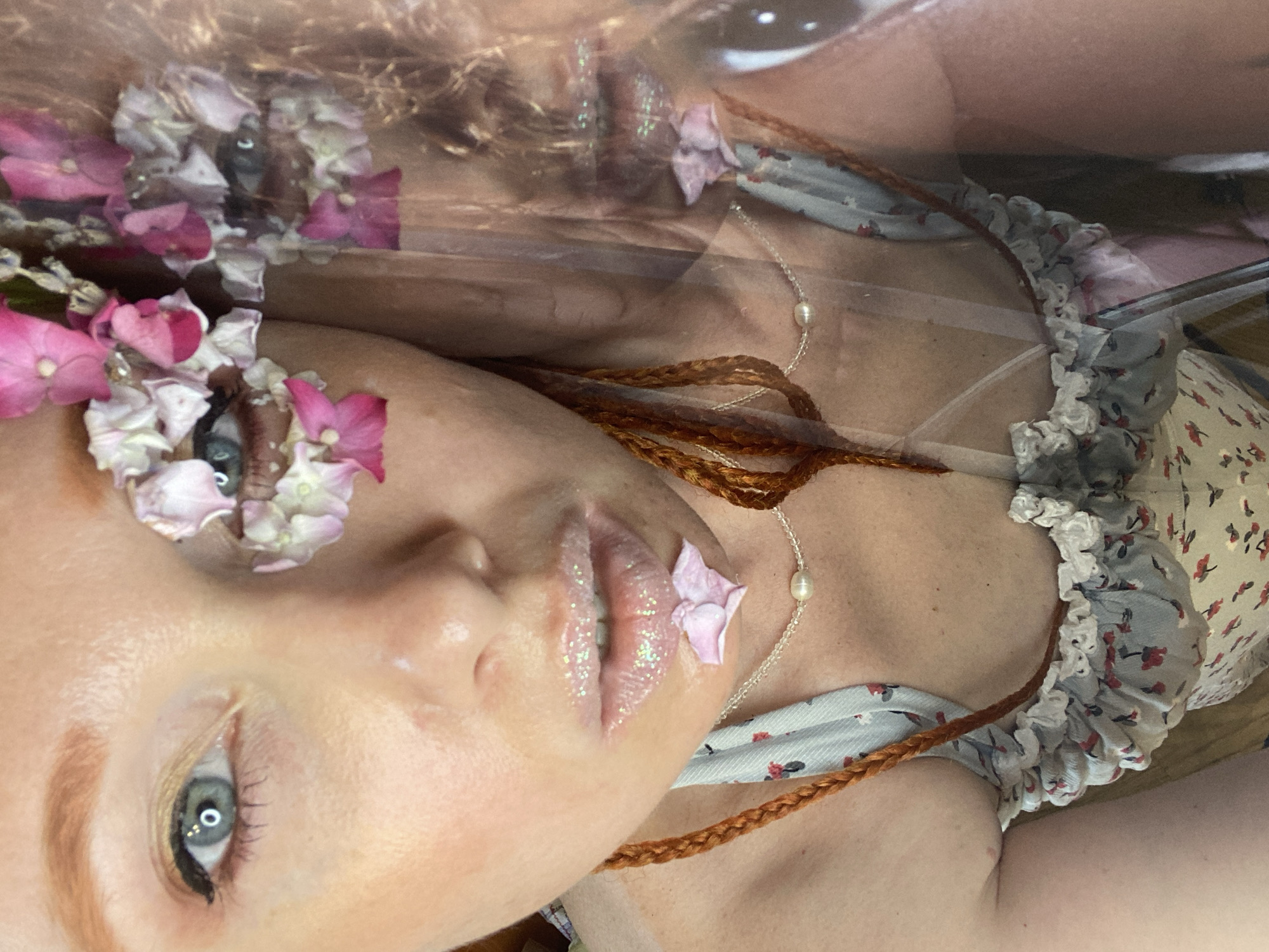 The image is a close-up, highly stylized self-portrait of a woman. She has light skin, red hair styled in braids, and blue eyes. Her face is adorned with pink and white flowers, with some covering her eyes. Her lips are covered in glittery lip gloss, and a single pink petal rests on her lower lip. She is wearing a light-colored, floral-patterned garment with ruffled edges and a delicate pearl necklace. The image has a double exposure effect, showing a slightly different, more transparent reflection of the woman's face and upper body. The overall aesthetic is ethereal and artistic. The image does not contain any information that would allow for its location to be determined in Sochi, Russia.