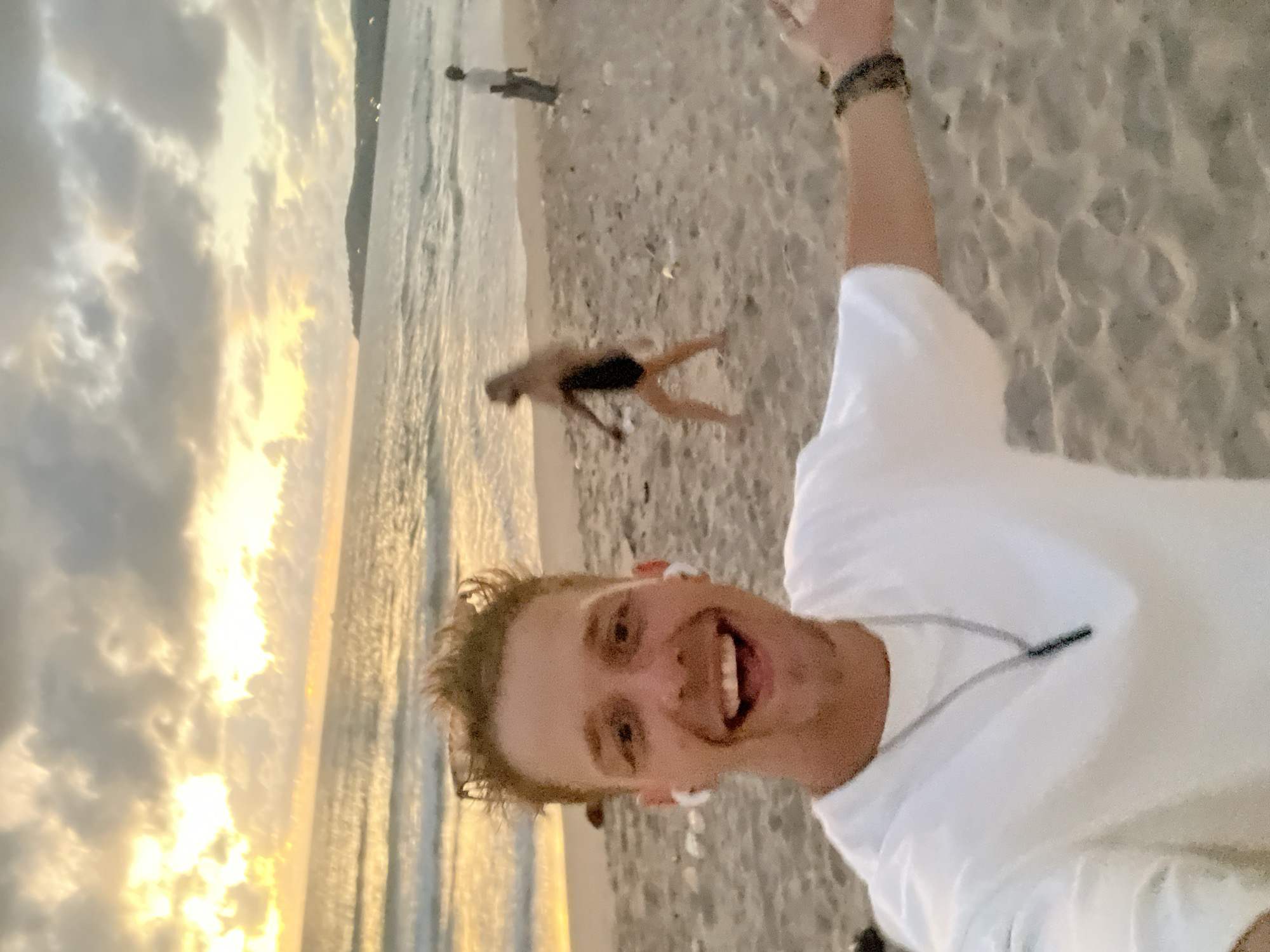 The image shows a selfie taken by a man on a beach at sunset. He is wearing a white t-shirt and has earbuds in. The man is smiling broadly, and his arm is raised. In the background, the ocean meets the sandy beach, with soft waves rolling in. The sky is filled with dramatic clouds, illuminated by the warm, golden light of the setting sun, creating a beautiful, atmospheric scene. A few other people are visible in the distance, walking along the shoreline. The overall mood is joyful and energetic, capturing a moment of happiness during a beach run or walk. The location is identified as Choeng Thale, Thailand, which is known for its beautiful beaches.