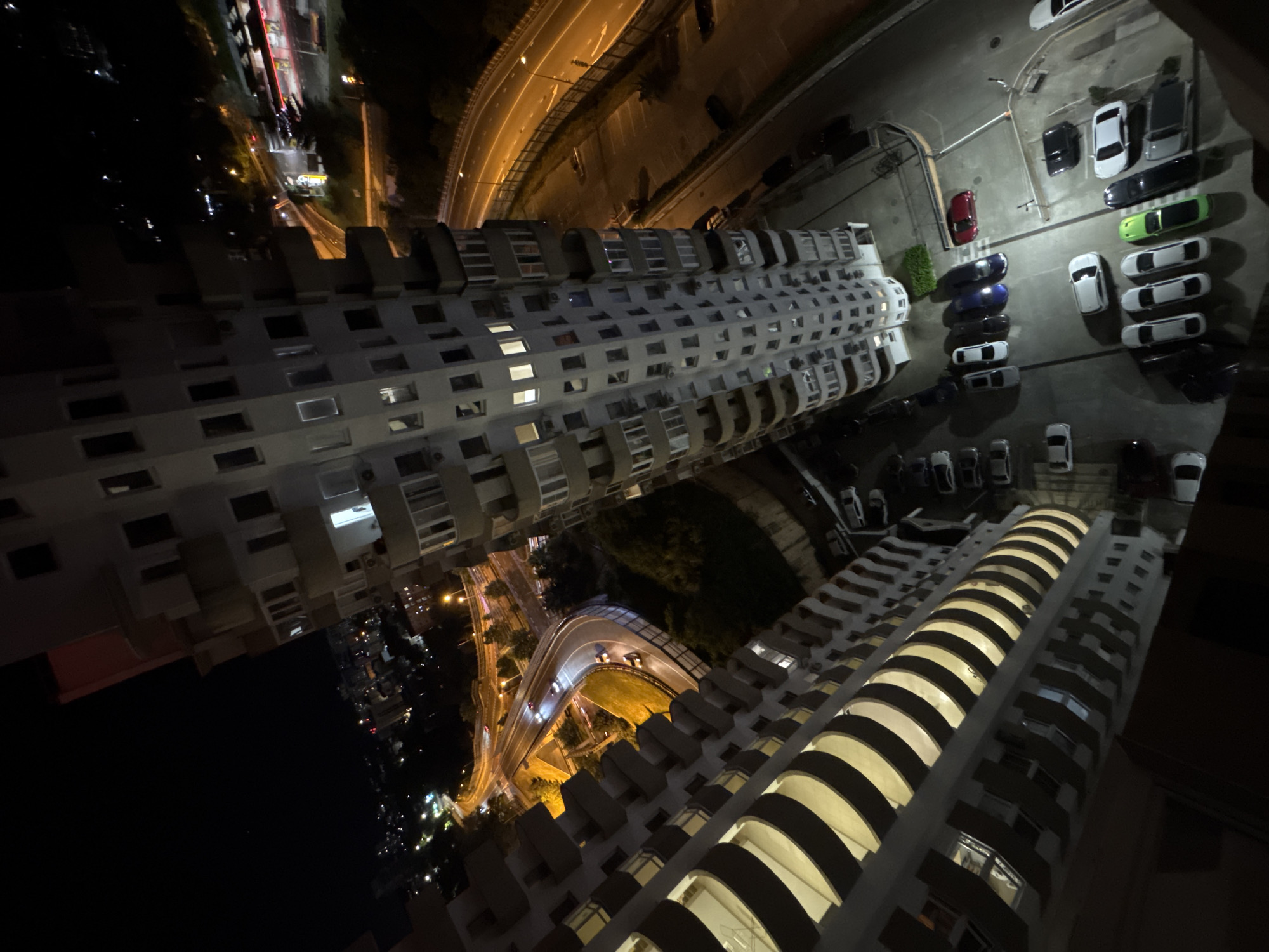 The image is a nighttime aerial view of a coastal city, likely Sochi, Russia, given the architectural style of the buildings and the presence of a highway. The scene is dominated by two tall, modern apartment buildings with curved balconies. The buildings are illuminated from within, with some windows showing lit interiors.
Below the buildings, a multi-lane highway curves through the landscape, brightly lit by streetlights. Cars are visible on the highway, some with their headlights on, indicating traffic. To the right of the highway, a parking lot is filled with various cars, including sedans, SUVs, and a bright red sports car. The surrounding area is a mix of urban development and greenery, with trees visible in the background. The overall impression is one of a bustling urban environment at night, with the city lights reflecting the vibrant life below.