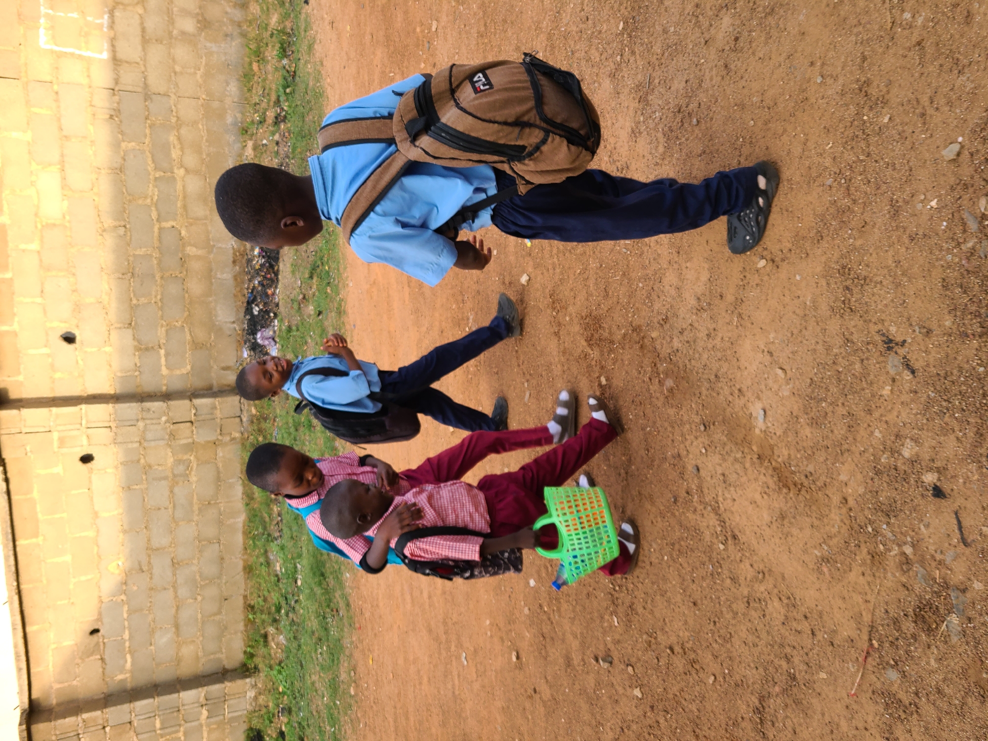 Three children in school uniforms are walking on a dirt road in Barakin Lerawi, Nigeria. The first child, wearing a light blue shirt and dark blue pants, has a brown backpack. The second child, wearing a light blue shirt and dark blue pants, also has a backpack. The third child, wearing a red and white checkered shirt and maroon pants, carries a green plastic basket. A brick wall is visible in the background, along with some grass. The road is sandy and there is some sparse vegetation.