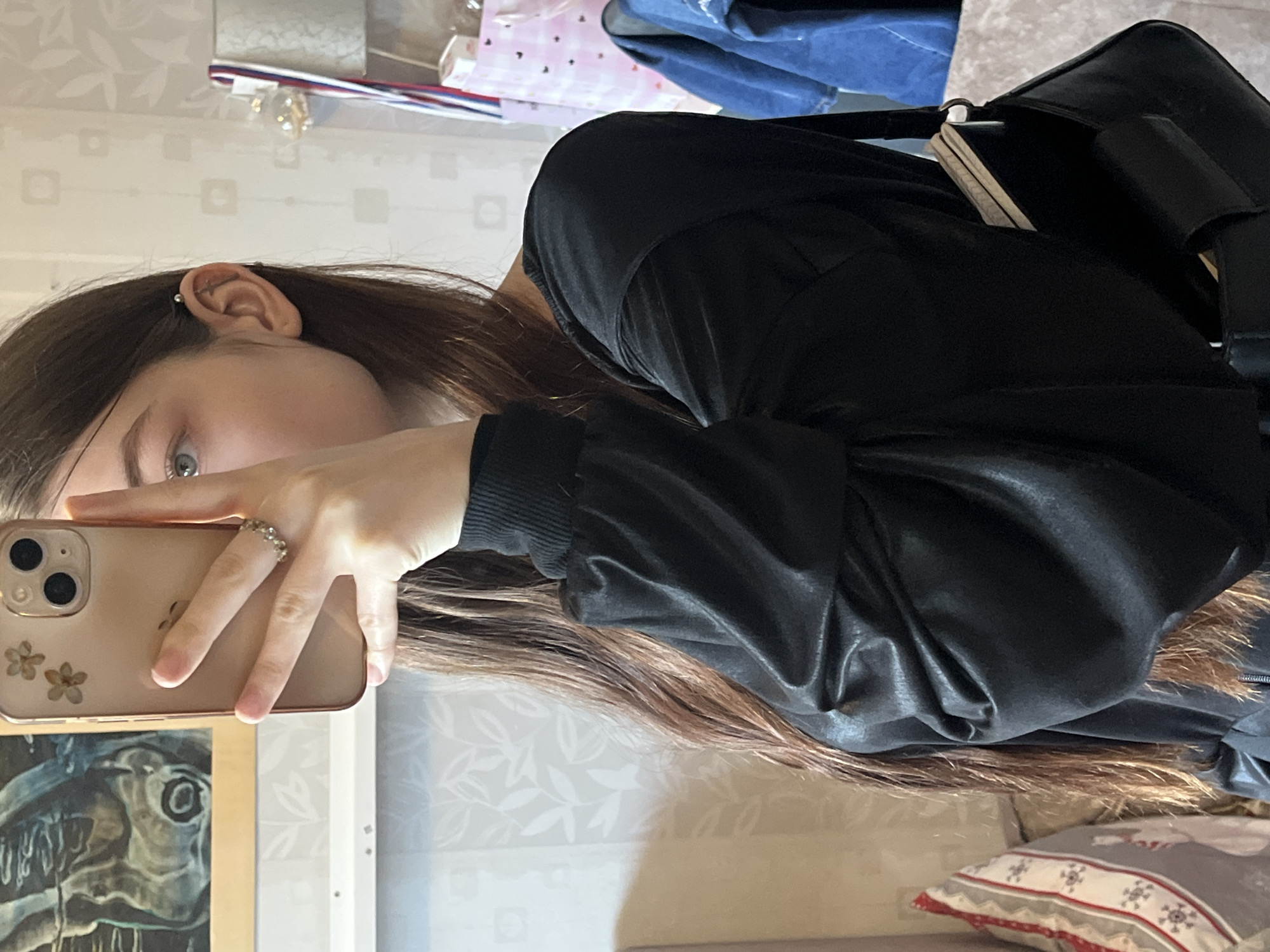The image, taken as a mirror selfie, shows a person, likely female, indoors. The subject is seen from the chest up, her face partially obscured by a light-colored smartphone held in her hand. She has long, dark brown hair and wears a black, long-sleeved top or jacket with ribbed cuffs, which appears to have a slightly shiny texture. Her visible left eye is light blue or grey, looking towards the camera. A bar-style piercing is visible on the upper helix of her ear.
Her hand, adorned with a silver-toned ring featuring small, shimmering elements, holds the smartphone. The phone's back is a light, possibly rose gold or champagne, color, showcasing two camera lenses and two decorative dried flowers embedded in the case.
The background reveals a light-colored wallpaper with a subtle, repeating leaf pattern. To the lower left, a framed artwork depicting a stylized, dark-toned fish or marine creature is reflected. To the upper right, a black bag or backpack is visible, with a light-colored notebook or papers partially sticking out. Above the bag, on a shelf or surface, are various items including ribbons with red, white, and blue stripes, and small shiny decorative objects. A blue fabric or garment is also visible behind the bag. In the bottom right corner, part of a decorative pillow with a red, white, and grey pattern, possibly snowflakes or stars, rests on a surface. The scene is brightly lit, suggesting it is daytime. No explicit text is visible.