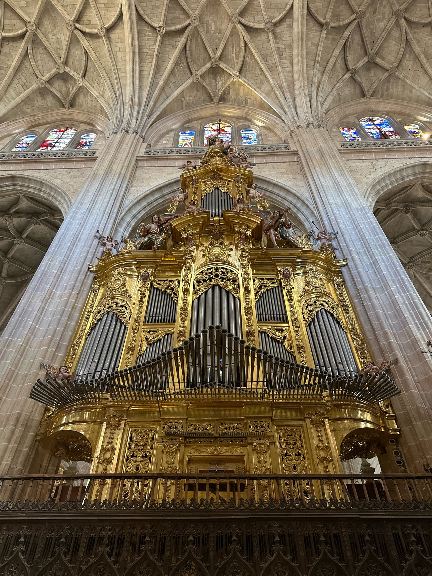 The image captures the ornate golden pipe organ within the Segovia Cathedral in La Lastrilla, Spain. The organ, a grand and intricate piece of craftsmanship, dominates the center of the frame. Its gleaming gold exterior is adorned with elaborate carvings and decorative elements, including angelic figures and facial motifs. Rows of metal pipes of varying sizes are prominently displayed, suggesting the instrument's powerful capabilities.
Above the organ, the soaring Gothic architecture of the cathedral is visible, characterized by ribbed vaults and pointed arches that ascend towards the heavens. Stained-glass windows with vibrant colors are set into the upper walls, allowing light to filter into the vast space. A wooden balcony, featuring decorative railings, extends from the organ's base, offering a vantage point to observe the instrument and the surrounding cathedral. The overall impression is one of immense scale, historical grandeur, and artistic splendor.