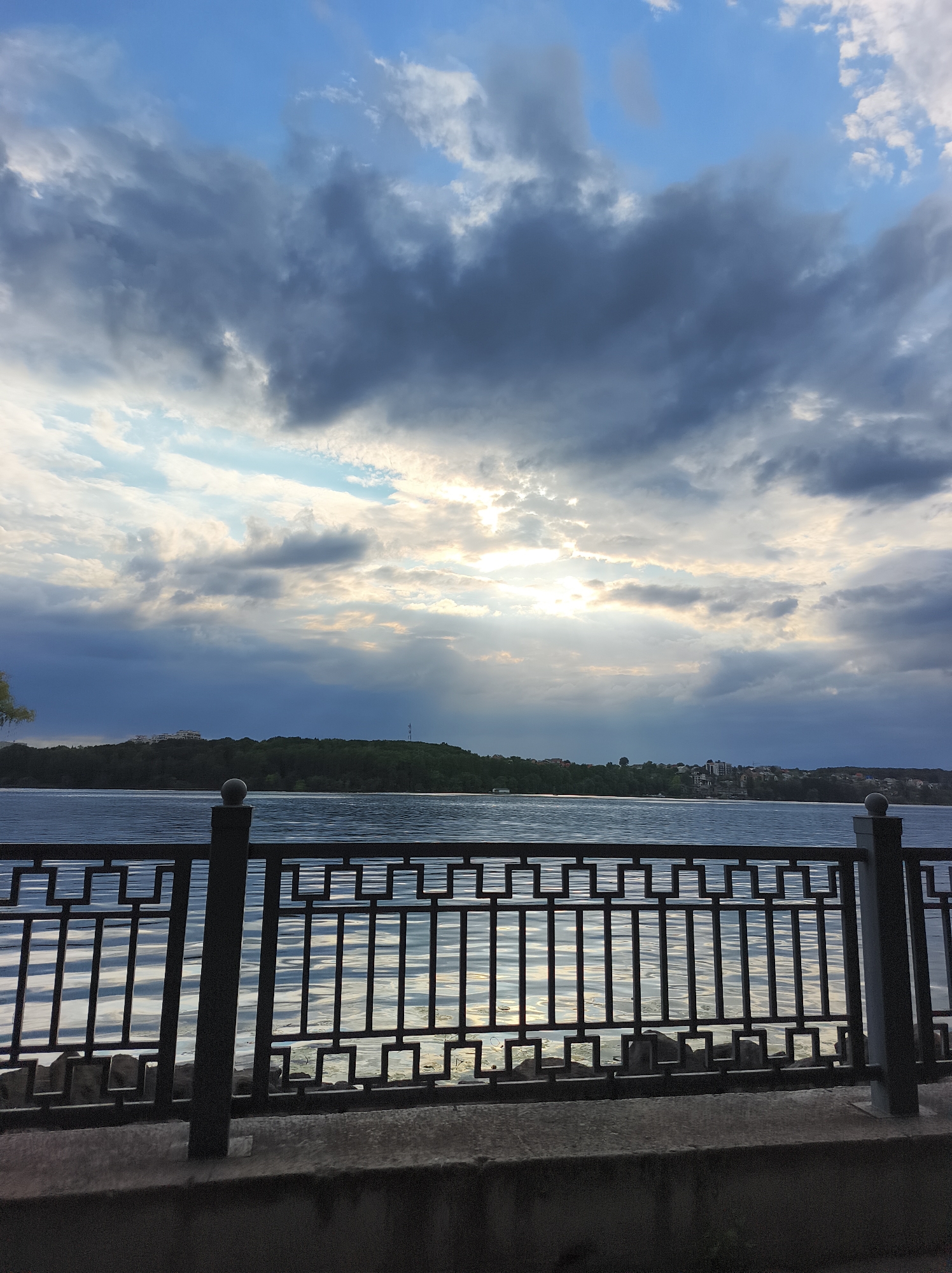 The image depicts a view of the Ternopil lake in Ternopil, Ukraine, during a cloudy sunset. The foreground features a dark metal railing with a geometric pattern, and a concrete walkway. Beyond the railing, the lake's surface reflects the sky, with ripples and light glinting on the water. In the background, a densely wooded hill rises, dotted with buildings, and a telecommunications tower is visible on the right. The sky is filled with dramatic, dark grey clouds, with patches of lighter blue sky and sunbeams breaking through, creating a moody and atmospheric scene. No people are present in the image.