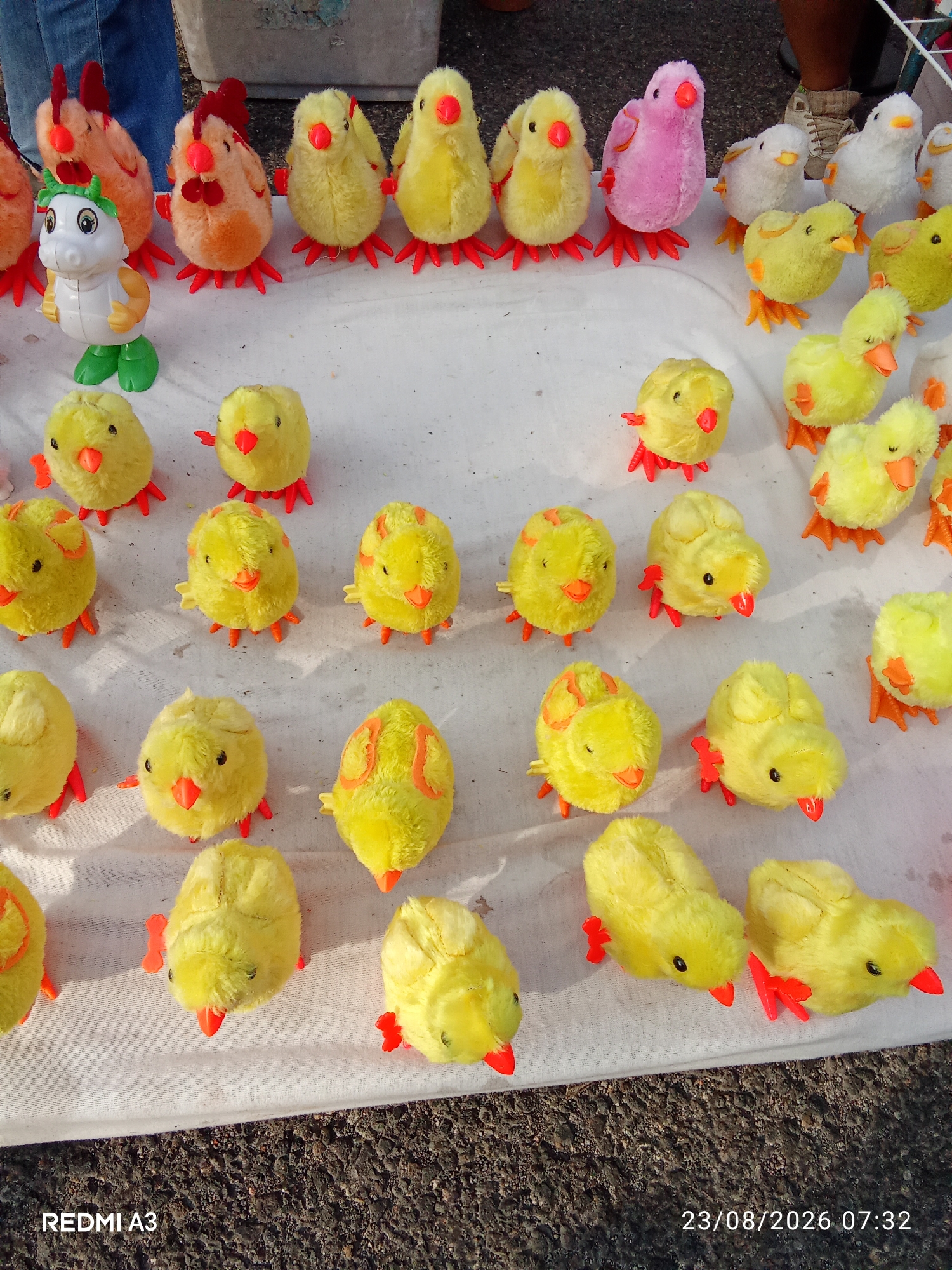 In a street setting in Grogol, Indonesia, a collection of plush toy chickens and chicks are displayed on a white cloth. The toys are varied in color, with orange, yellow, pink, and white chicks, and a few larger orange chickens with red combs. Some of the chicks appear to be wind-up toys, with visible red plastic feet. In the background, a person's legs in blue jeans are visible, as is a gray bin. The ground beneath the cloth appears to be asphalt. The image is dated August 23, 2026, at 07:32.