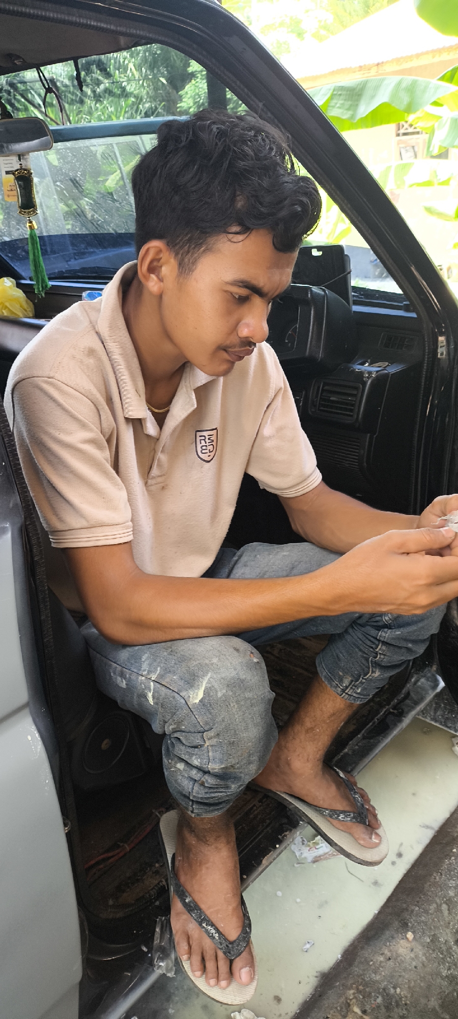 An overhead shot shows a young Indonesian man with dark, curly hair and a mustache sitting in the open driver's side door of a vehicle. He is wearing a tan polo shirt with a crest on the left breast and faded blue jeans speckled with white paint. His bare feet are clad in black flip-flops. His left foot rests on the vehicle's floorboard, which appears to be dirty and wet, while his right foot is propped up slightly on the exterior of the vehicle. He is looking down at his hands, which are holding a small, white, shredded object, possibly paper. The interior of the vehicle is dark, with black dashboard components visible. The exterior background consists of lush green foliage and the suggestion of buildings, indicating a tropical environment.