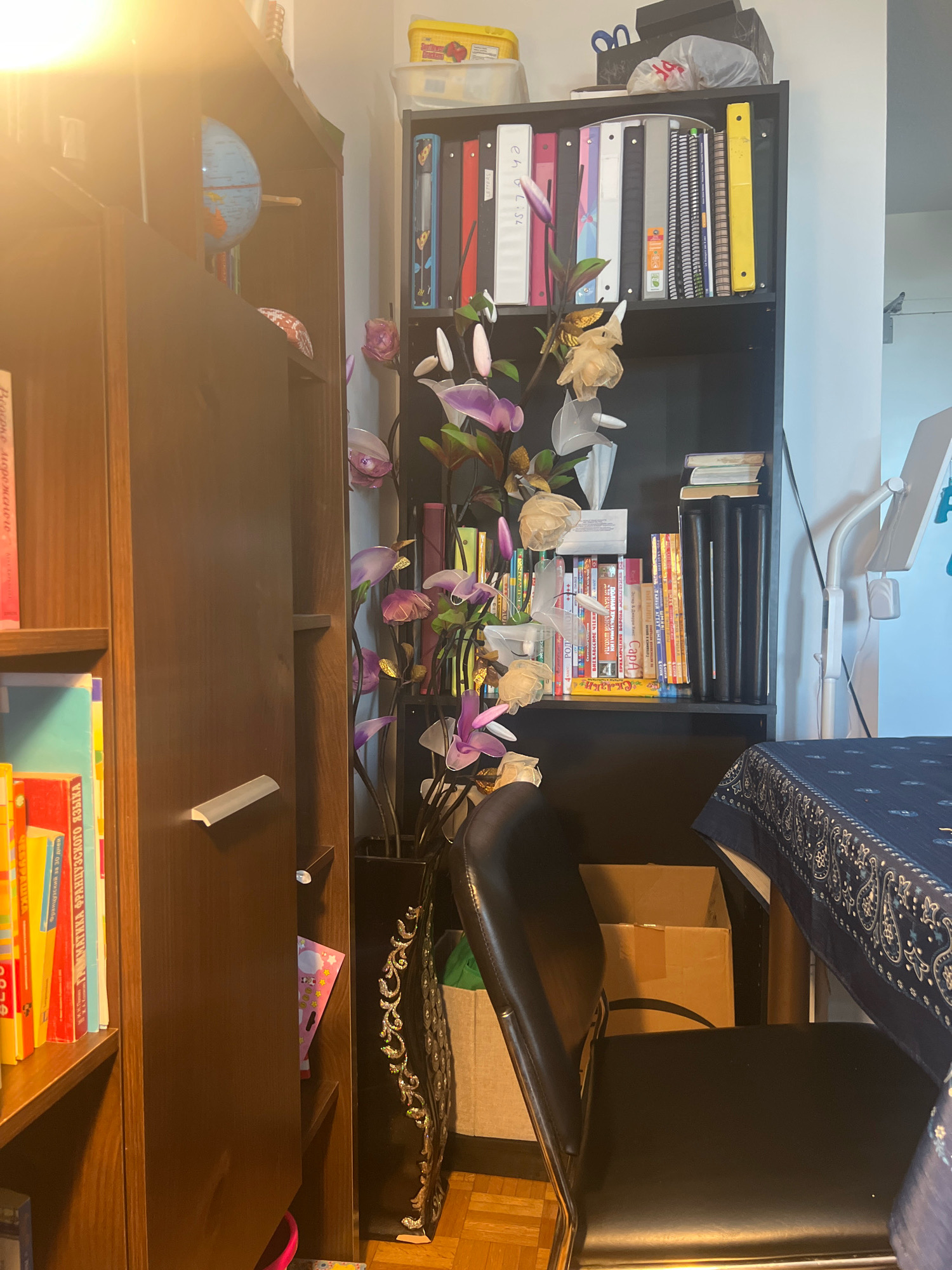 This is a medium shot, eye-level interior scene of a room, likely a study or living area, filled with books and decorative items. The lighting is warm and appears to be coming from the left, casting a bright glow on the left side of the image.
On the left, a tall, dark wood bookshelf is filled with books, some of which have colorful spines. A small globe is visible on the top shelf of this bookcase. To the right of this bookshelf is a black shelving unit packed with various binders, notebooks, and books, arranged vertically. Among these items, a decorative artificial plant with purple and cream flowers stands tall, adding a touch of nature to the space. On top of the black shelving unit, there are some stacked boxes and bags, including what appears to be a yellow container of food.
In the foreground, a dark leather chair with a chrome frame is positioned in front of a table covered with a dark blue patterned cloth. A cardboard box is visible behind the chair. The flooring appears to be wood laminate.
In the background, a white floor lamp with a tablet holder is visible on the far right. The overall environment suggests a home setting, possibly within the city of Markham, Canada, characterized by its organized clutter of personal belongings and functional furniture.