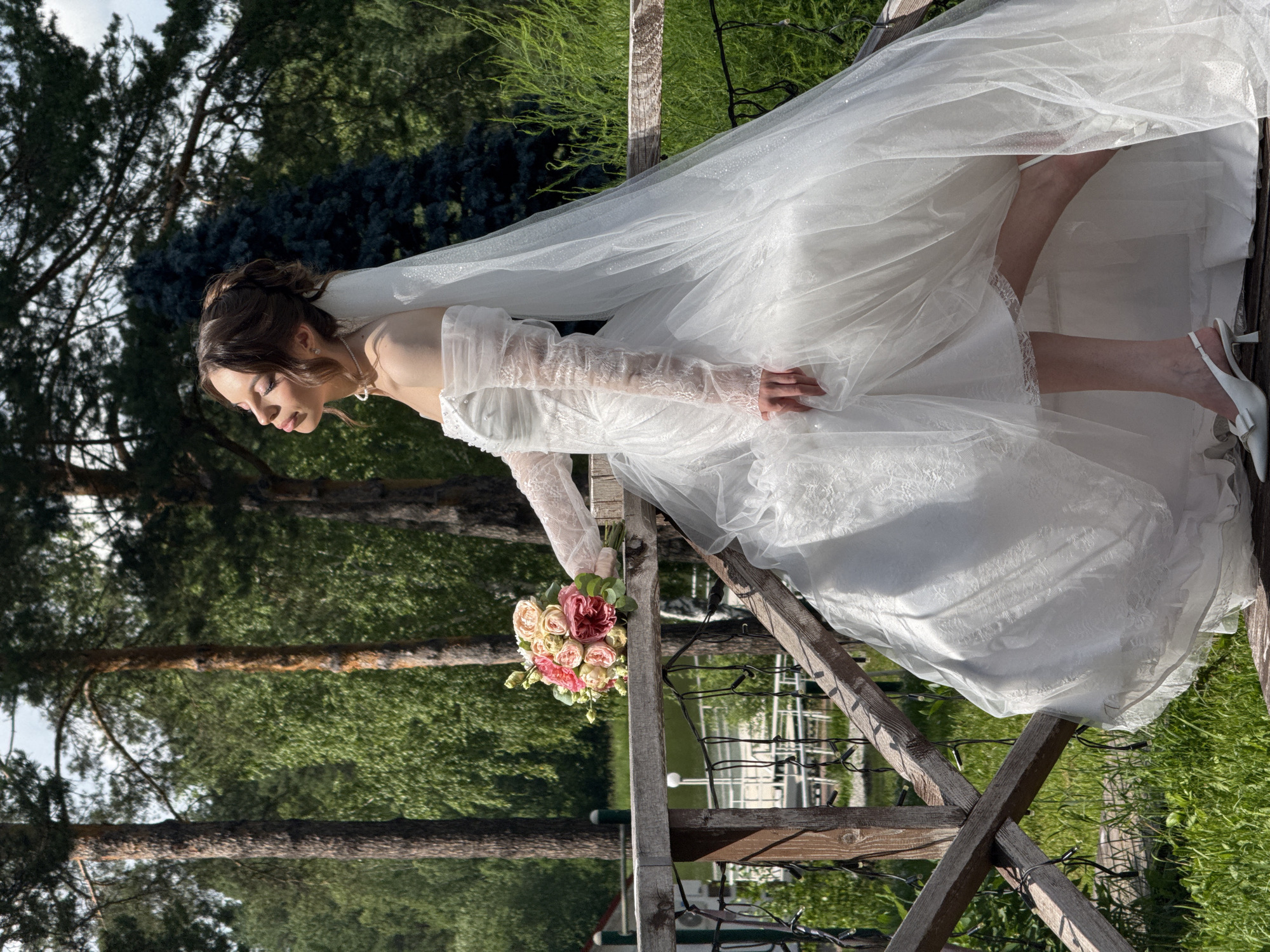 This is a vertical, outdoor photograph of a bride in a white wedding dress with a veil and long lace sleeves. She is holding a bouquet of pink and white roses. The bride is looking down and to her left with a gentle smile, suggesting a moment of reflection or happiness. She is wearing white heeled shoes.
The scene is set outdoors with lush green trees and foliage in the background. The bride is positioned on a wooden structure, possibly a gazebo or a decorative arbor, with wooden beams and railings visible. String lights are draped around some of the wooden elements. The lighting suggests it is daytime, likely sunny, as indicated by the shadows and highlights on the dress and veil, which has a subtle sparkle.
The location context places this scene in Votkinsk, Russia. The overall mood is romantic and serene, capturing a tranquil moment of a bride on her wedding day amidst nature. There is no visible text in the image.
