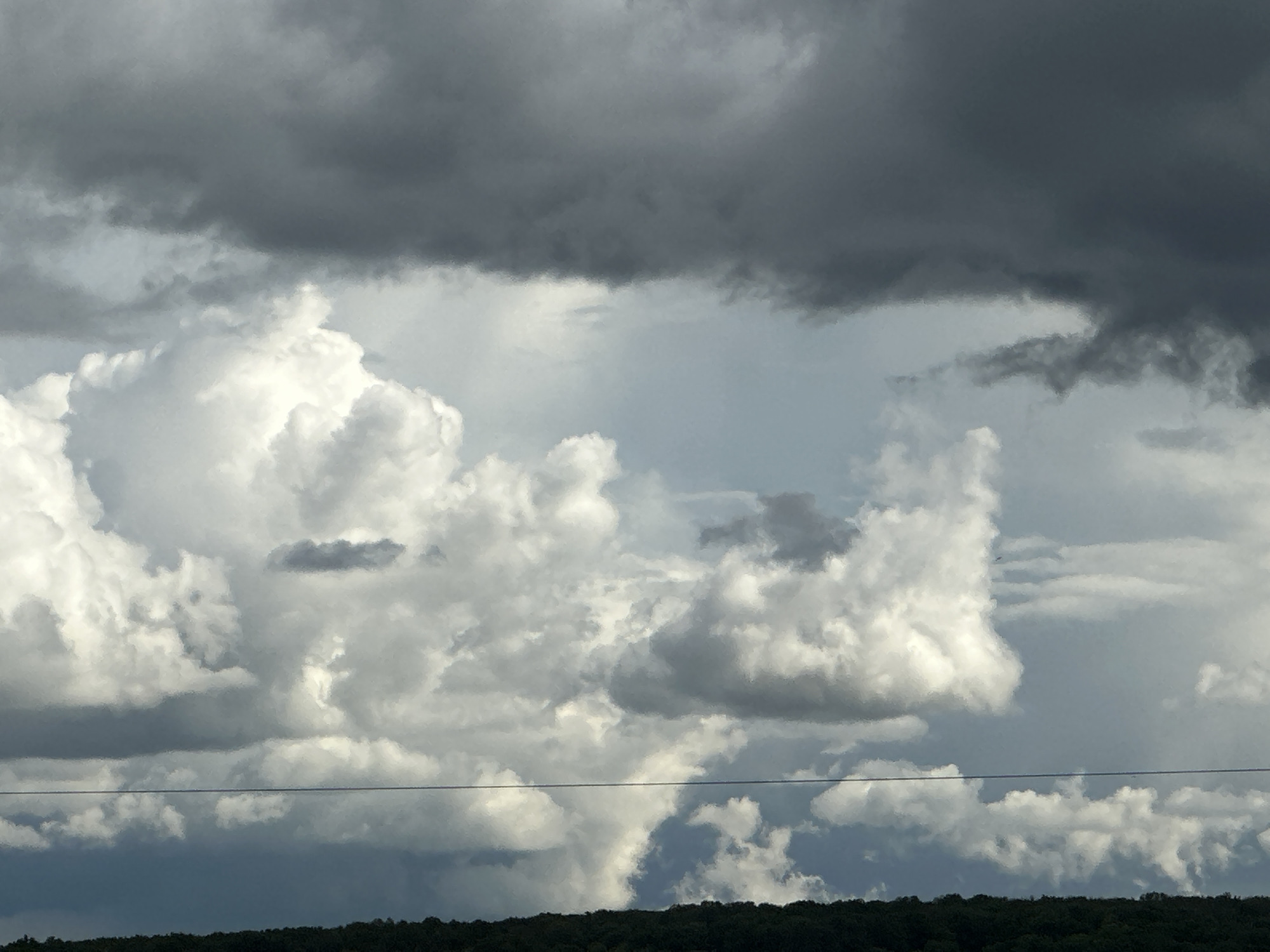 The image depicts a sky filled with a mix of cumulus and cumulonimbus clouds. Dark, ominous storm clouds dominate the upper portion of the frame, contrasting with the brighter, white cumulus clouds below. These cumulus clouds are puffy and scattered, with varying degrees of light and shadow. A thin, dark treeline is visible at the very bottom of the image, suggesting a landscape beneath the sky. A power line cuts horizontally across the middle of the frame. The location is Unknown, Unknown.