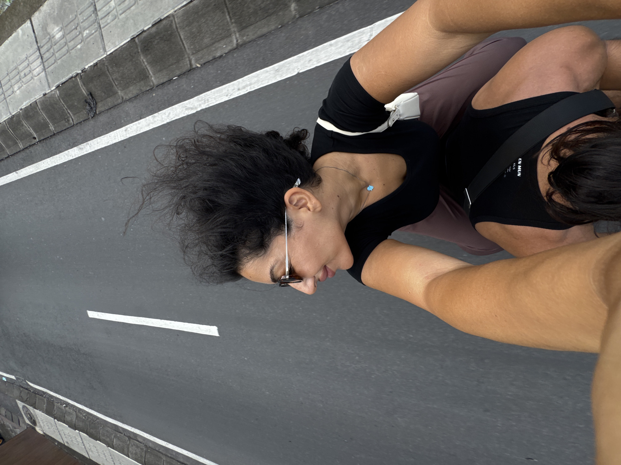 This dynamic, high-angle shot captures two or possibly three individuals traveling, likely on a scooter or motorcycle, in North Kuta, Indonesia. The perspective suggests a selfie taken by someone's outstretched arm.
In the foreground, a tanned arm and shoulder, belonging to the person holding the camera, are visible on the right. Directly in front of the camera, a person with dark, curly hair is seen in profile, looking slightly ahead and downwards, displaying a relaxed or subtly smiling expression. She wears a black short-sleeved top, sunglasses perched on her head, and a delicate light blue flower-shaped necklace. A small white shoulder bag rests on her left side. Behind her, another person's back is visible, wearing a black tank top with a broad black strap across their shoulder. Partial text "EN MUN" and "XS/S" can be read on this strap. The lower part of their legs or thighs, also tanned, are seen behind the second person.
The subjects are positioned over an asphalt road featuring a crisp white solid line. To the left, a textured concrete sidewalk or curb with distinct block patterns runs parallel to the road. The lighting indicates it is daytime with good visibility. The overall scene conveys a sense of active travel through an urban or semi-urban environment in North Kuta.