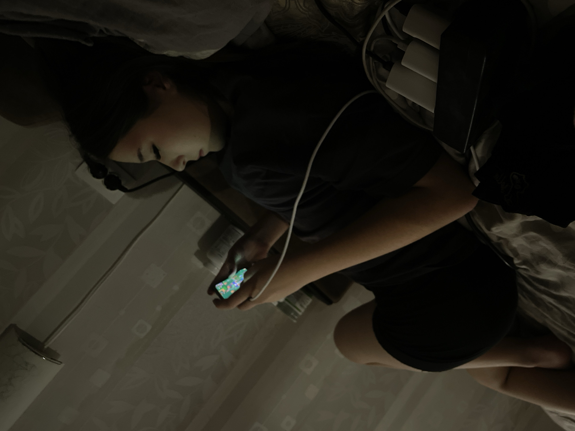 An image, rotated clockwise, depicts a young female lying down in a dimly lit indoor setting, likely a bedroom or living room, characteristic of a private residence in Saint Petersburg, Russia. The scene suggests nighttime or very low ambient light.
The subject, seen in profile, has dark hair and is wearing a dark short-sleeved t-shirt and dark shorts. Her face is gently illuminated by the bright, colorful screen of a smartphone held in her right hand, which appears to be connected to a white charging cable. Her left hand is also visible, resting near the phone. Her gaze is directed downwards at the screen, indicating focus.
Around her head, several white charging adapters are visible, plugged into a black power strip or multi-port charger, with various cables tangled nearby. She is lying on a surface covered with rumpled fabric, suggesting a bed or couch. The wall in the background features a subtle, light-colored floral or leafy wallpaper pattern. A white cylindrical object, possibly a lamp base, is partially visible in the bottom-left corner of the frame. The primary activity is the individual using her smartphone while it is charging. No explicit emotions are discernable, and there is no visible text in the image.