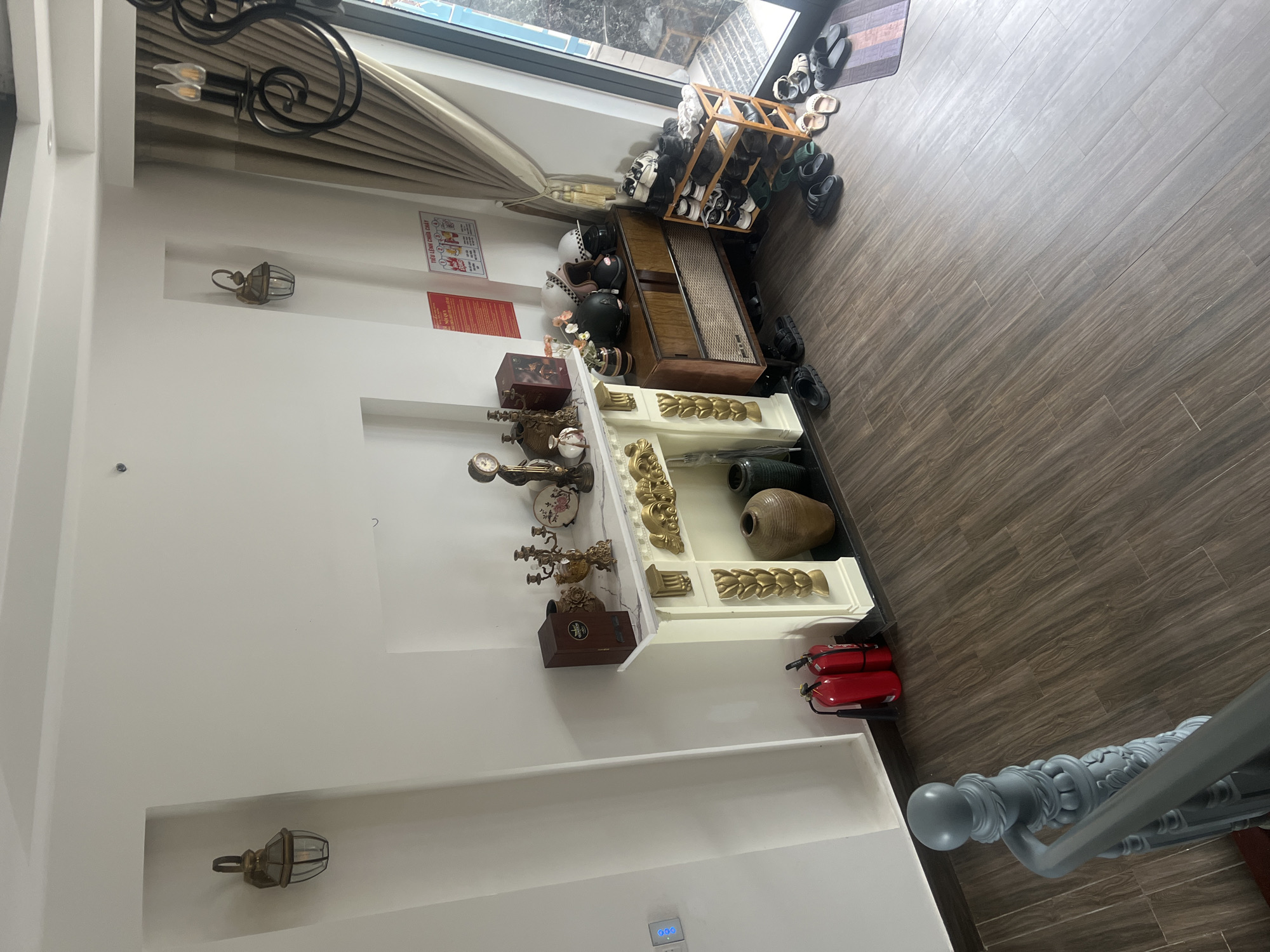 The image shows the interior of a home, likely in Đà Nẵng, Vietnam. The space features tiled flooring, a white wall with recessed shelves containing decorative items and a mounted lantern-style light fixture. A display stand with various antique-style objects, including vases, clocks, and decorative gold pieces, is against the wall. Near the entrance, there's a shoe rack, helmets, and a wooden cabinet with a speaker grill on the front. Two red fire extinguishers are on the floor next to the display stand. The scene suggests a decorated, lived-in space with a mix of functional and decorative elements.