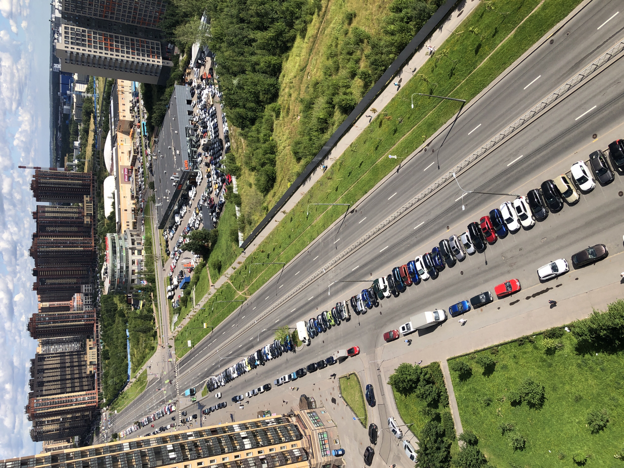 An aerial, medium shot captures a sunlit street in a modern urban area with high-rise buildings and a large commercial complex on the right. The street runs diagonally from the left foreground to the upper right background. On the left, a tall, beige residential building dominates the foreground. Across the street, numerous cars are parked in a long line along the sidewalk. The street itself has multiple lanes and is busy with traffic. A large, dark-roofed commercial building, identified by a "LADA" sign, is visible in the middle ground on the right, with a large lot filled with cars. Further back, a lighter-colored commercial building with a rounded facade and green landscaping is seen. The background features more residential buildings and some industrial structures. The sky is partly cloudy with bright sunlight casting shadows. People are visible walking on the sidewalks and in the parking lot.