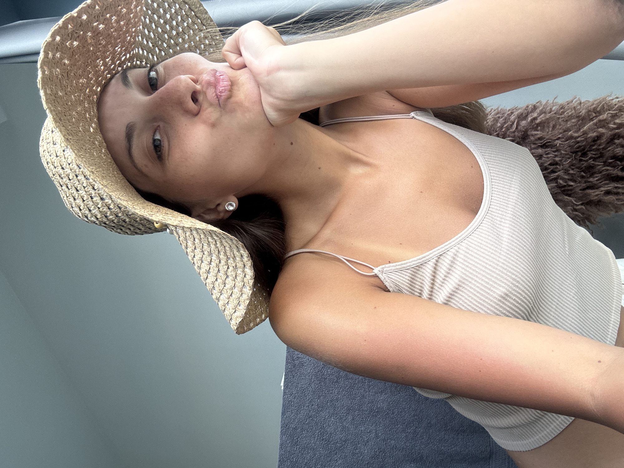 The image shows a young woman in a playful pose, rotated counter-clockwise such that her head is positioned on the left side of the frame. She is wearing a wide-brimmed straw hat that covers most of her head, and a light-colored, possibly beige or off-white, ribbed tank top with thin spaghetti straps. Her dark hair is visible beneath the hat and flowing towards the right.
Her face is turned upwards towards the viewer, displaying a distinct pouting or "fish face" expression with puckered lips. Her left hand is pressed against her cheek and chin, gently pushing up the skin, which emphasizes the playful pout. A small, sparkling diamond stud earring is visible in her right ear. Her skin tone appears lightly tanned, with a noticeable reddish tint on her left arm, potentially indicating recent sun exposure.
The background on the left side consists of a plain, muted blue-grey surface, possibly an interior wall or panel. To the right, behind her shoulder and arm, a textured, fluffy brown material is visible, which could be a blanket, cushion, or part of a piece of furniture. The lighting is bright and appears natural, highlighting her features clearly. There is no visible text in the image. The context for the location is Kuala Lumpur, Malaysia.