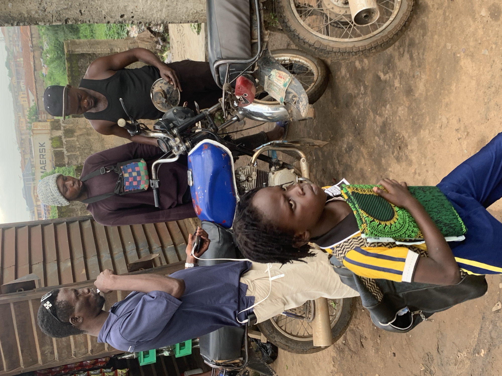 In Shagamu, Nigeria, a street scene unfolds as three men and a young child stand near two motorcycles. The foreground is occupied by a young child with dark, dreadlocked hair, wearing a vibrant yellow, black, and blue striped shirt and blue pants. They carry a black backpack and clutch a green fabric with a circular pattern. To the left, a man in a navy blue polo shirt and khaki pants, wearing sunglasses pushed up on his head, appears to be chewing on something and holding a phone with a charger cord extending to a power bank in his pocket. Behind him, a man in a maroon long-sleeved shirt and a patterned crossbody bag stands by a blue and silver motorcycle. To the right, a man in a black tank top and black cap holds a motorcycle part, possibly a headlight assembly. The background reveals a rustic environment with buildings, greenery, and a shipping container with the word "MAERSK" visible. The ground is unpaved, suggesting a typical street or market setting in the city.