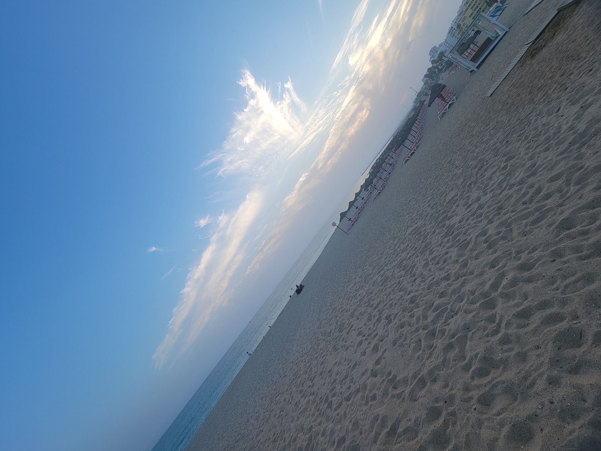 The image depicts a wide, sandy beach in Quarteira, Portugal, likely during the late afternoon or early evening. The sand is textured with footprints and ripples, suggesting it has been recently disturbed. In the distance, rows of unoccupied beach chairs and thatched umbrellas stretch along the shoreline, indicating the beach is either preparing for the evening or is in a less crowded season. The calm sea is visible on the left, meeting a hazy horizon. Above, the sky is a clear, bright blue, transitioning to softer hues near the horizon where wispy white clouds are illuminated by the setting sun. A few scattered individuals are present on the beach, with one person lying on a beach chair in the middle ground and another person visible near the water's edge. The overall scene is serene and tranquil.