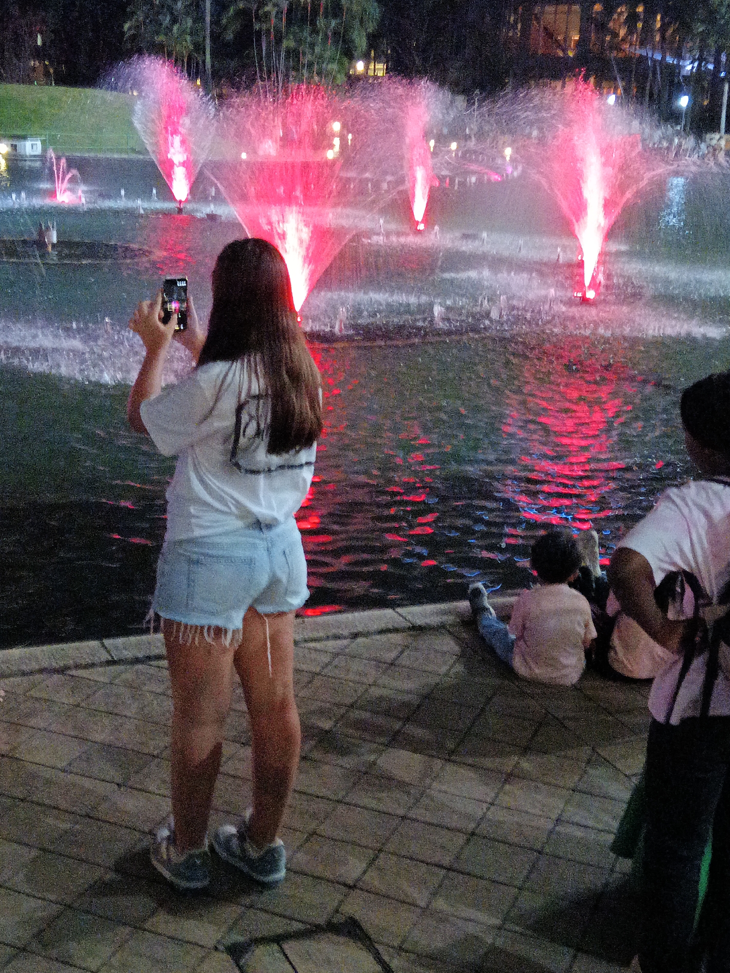 A woman stands facing away from the camera, holding a mobile phone in her hands, as if taking a photo or video. She is wearing a white t-shirt and light blue denim shorts. In front of her, a large fountain sprays water illuminated by pink lights. The water reflects the pink light onto the surface of the fountain's basin. To her right, two children are sitting on the ground, facing the fountain. The environment appears to be an outdoor plaza or park, possibly in Kuala Lumpur, Malaysia, given the presence of palm trees and urban lighting in the background. The ground is paved with interlocking bricks.