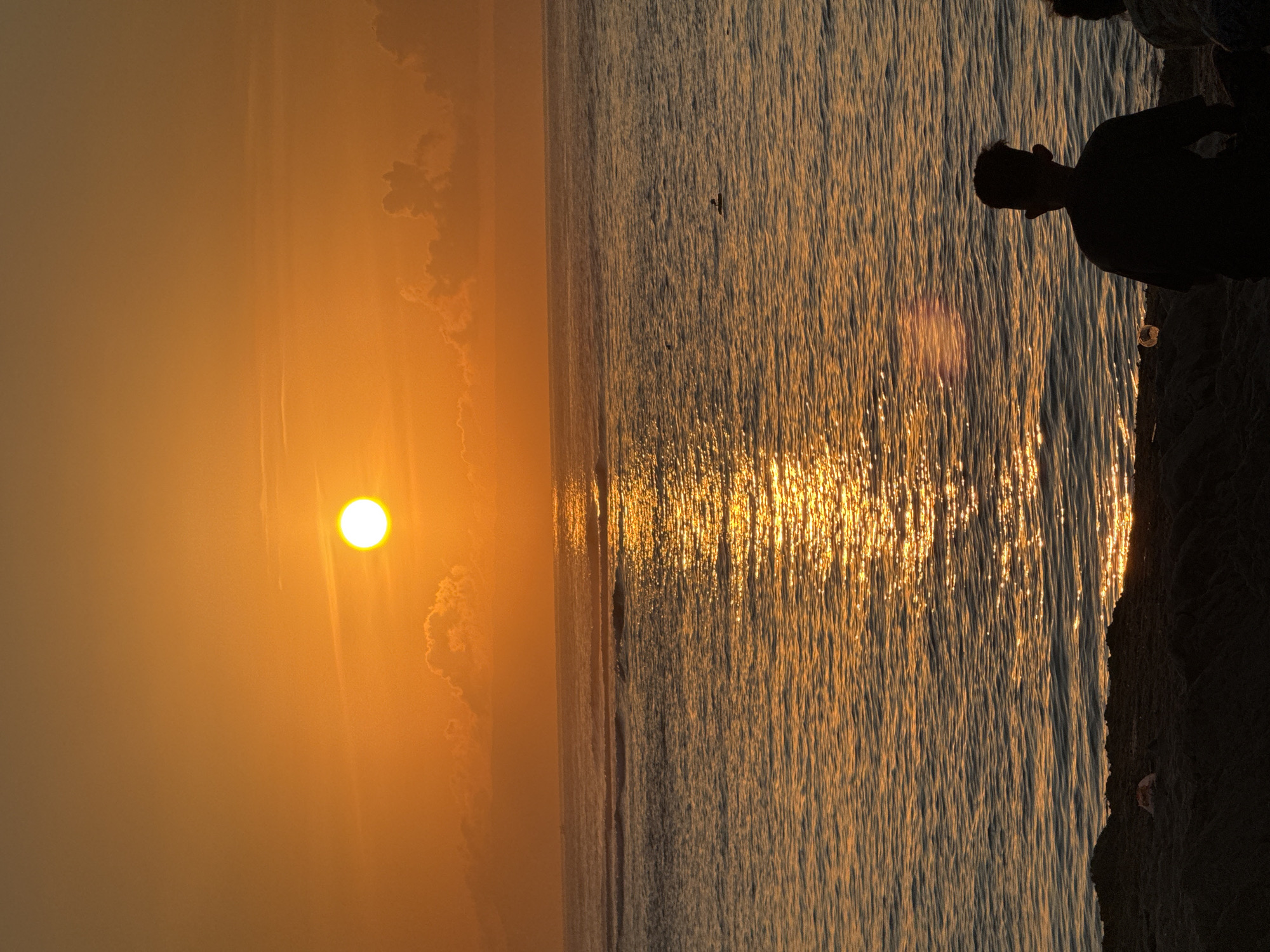The image shows a sunset over the ocean, likely taken at the southern end of Kuta Selatan, Indonesia. The sun, a bright white-yellow orb, is partially obscured by thin clouds. Reflections of sunlight glisten across the surface of the water. A person's silhouette is visible on the right side of the frame, near what appears to be a rocky shoreline.