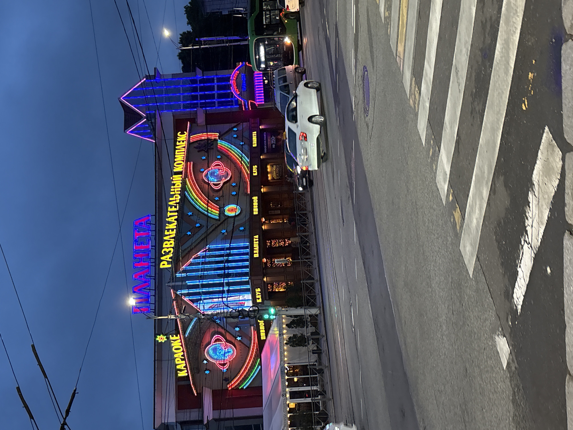 This image depicts the "Planeta Entertainment Complex" located at Chernyakhovskogo St, 28, Kaliningrad, Kaliningrad Oblast, Russia, during dusk or night hours. The two-story building is prominently illuminated with numerous neon lights. The main sign at the top reads "ПЛАНЕТА" (PLANETA) in pink neon, with "РАЗВЛЕКАТЕЛЬНЫЙ КОМПЛЕКС" (ENTERTAINMENT COMPLEX) in yellow neon directly below it. A "КАРАОКЕ" (KARAOKE) sign in yellow neon is visible on the left side of the facade. The building's exterior features a large neon art installation of a planet with concentric rings, encircled by a rainbow-colored arc and multiple stars. A section on the right side of the building exhibits vertical blue neon stripes, simulating windows, topped by a magenta-outlined roof structure. At street level, signs for "ПИВНОЙ КЛУБ" (BEER CLUB) and "КЛУБ ПЛАНЕТА" (CLUB PLANETA) are discernible. To the left of the entrance, a large white outdoor tent structure, labeled "ВЕРАНДА ПЛАНЕТА" (VERANDA PLANETA), occupies the sidewalk.
In the foreground, a multi-lane asphalt road with white lane markings and a partial pedestrian crosswalk (zebra stripes) on the lower right is visible. Traffic includes a white sedan (likely a Chevrolet Cobalt) moving right to left in the middle lane, a gray minivan behind it, and a dark sedan partially visible on the far left. A green public transit bus is also moving right to left on the right side of the road. Overhead wires, indicative of trolleybus or tram lines, stretch across the dark blue sky. A streetlight pole stands in front of the entertainment complex, and a green traffic signal is visible on the left. A metal fence borders the sidewalk in front of the building. No distinct individuals are clearly visible or identifiable within the frame.