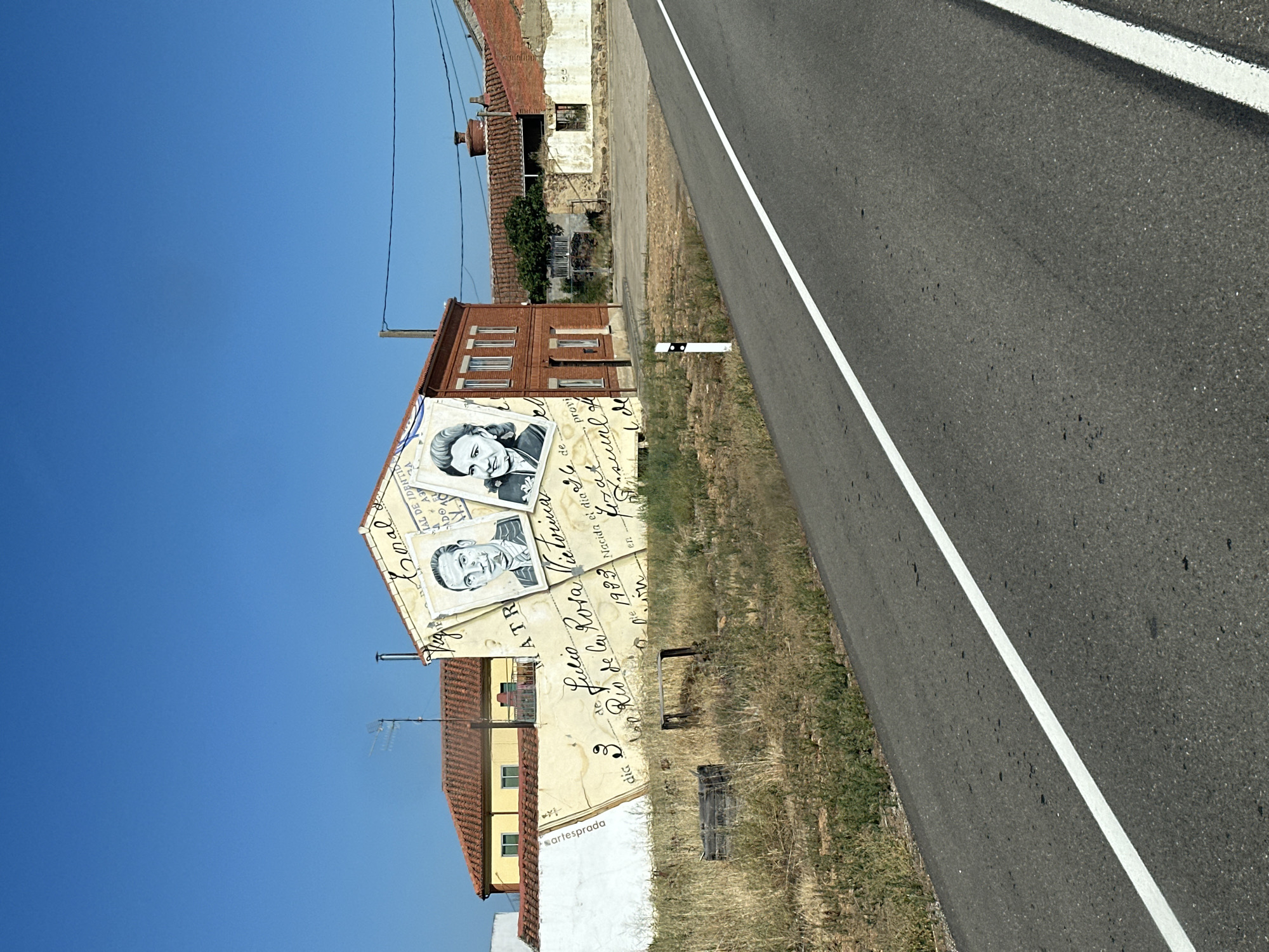 In Toral de Fondo, Spain, a mural is painted on the side of a building. The mural depicts a large, yellowed envelope with cursive writing and two portraits of individuals. The building itself is red brick with white-trimmed windows and a sloping tiled roof. The mural covers the side of the building, extending upwards to the roofline. Adjacent to this building is another structure with a red tiled roof, partially obscured. The foreground features a paved road with a white line marking its center, bordered by dry, grassy vegetation. The sky above is a clear, vibrant blue.
