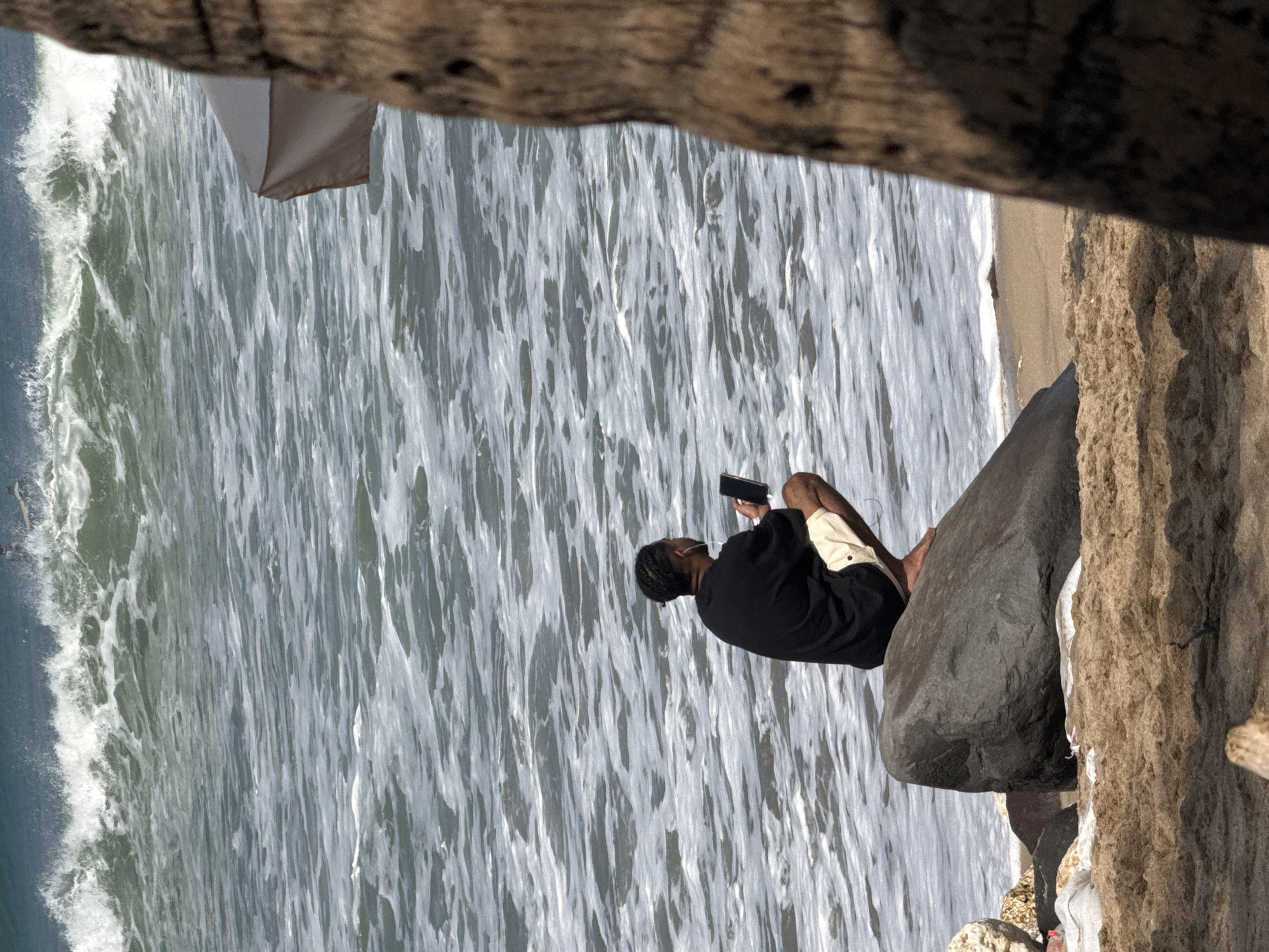 A person wearing a black t-shirt and light-colored shorts is squatting on a large rock on a sandy beach. The person is holding a black object, possibly a phone, in their right hand and is wearing white earbuds. In the background, the ocean has large waves crashing, with white foam visible. A partially visible beach umbrella and the trunk of a palm tree are to the right of the frame. The location is Kuta, Indonesia.