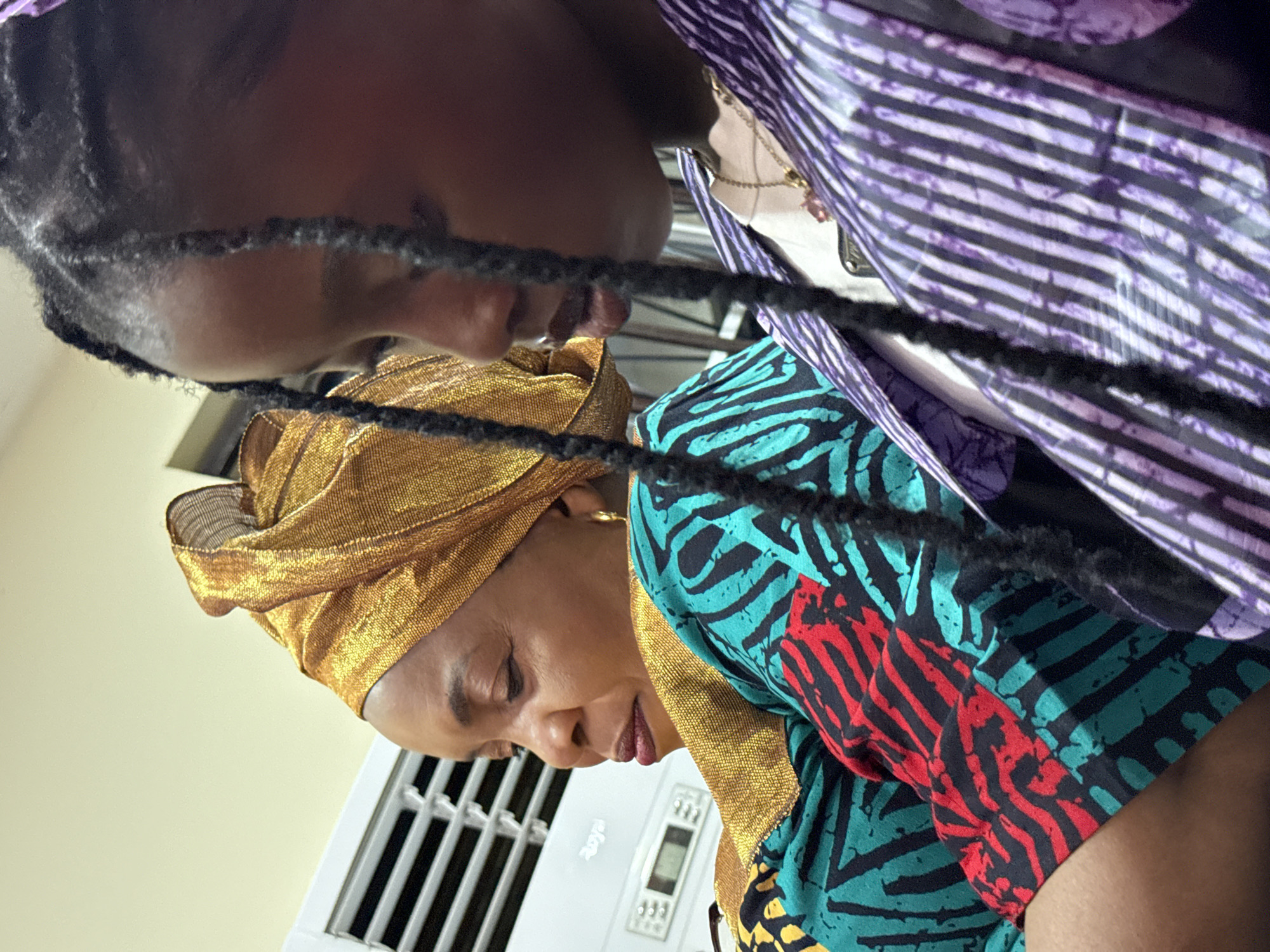 In Lugbe, Nigeria, two females are captured in a close-up, side-profile shot. The female on the left wears a striking gold headwrap and a vibrantly patterned top in shades of teal, red, and black. Her gaze is directed downwards, her expression thoughtful. To her right, a younger female is visible, her dark braided hair framing her face. She wears a purple and white striped garment, with a delicate gold necklace adorned with a pink pendant visible at her neck. The background is subtly illuminated, suggesting an indoor setting. An air conditioning unit is partially visible behind the female on the left, its white surface contrasting with the warm tones of the room.