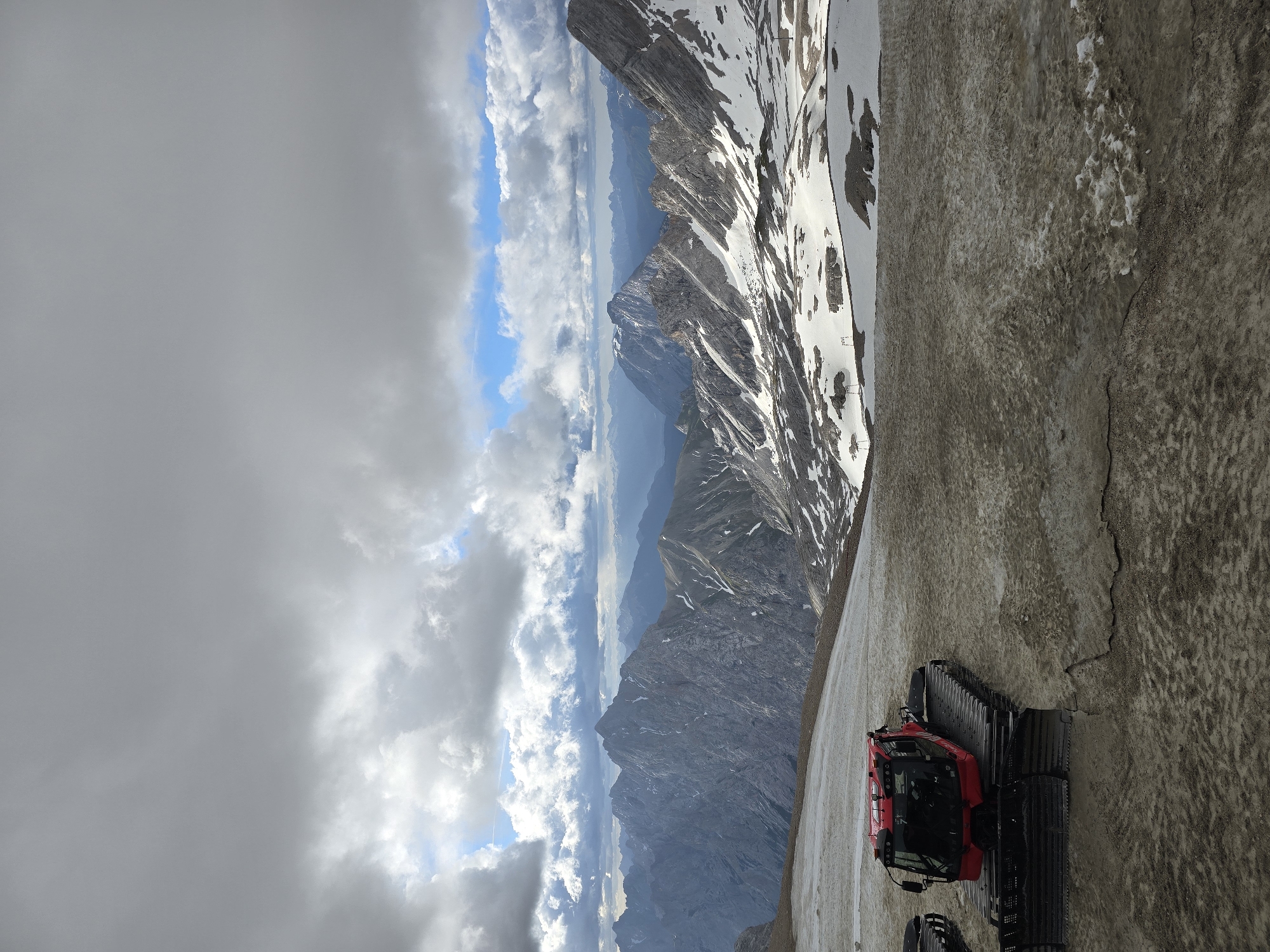 A red snow groomer is parked on a muddy, snow-covered slope. Behind it, a vast mountain range stretches into the distance under a cloudy sky. The mountains are rugged with patches of snow clinging to their slopes and peaks. The foreground shows a mixture of dirt and melting snow, indicating a transitional season. The scene is set in a high-altitude alpine environment, likely within the Bavarian Alps near Garmisch-Partenkirchen, Germany, given the distinctive mountain formations. There are no people visible in the image.