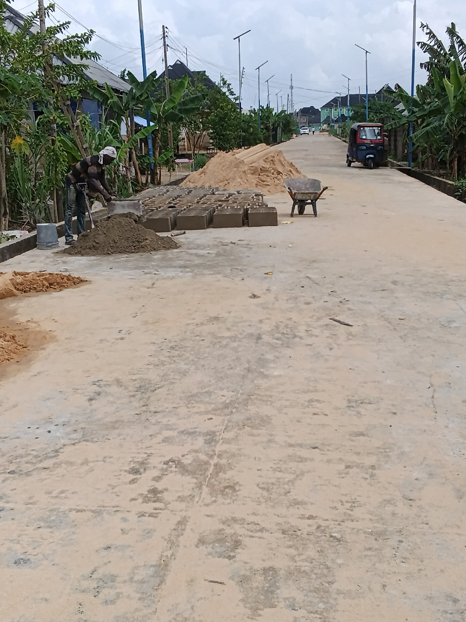 A man, wearing a striped shirt, dark pants, and a white headscarf, is making bricks on a paved road. He is using a shovel to mix sand and appears to be forming a brick. In the foreground, there are stacks of pre-made bricks and a pile of sand. A wheelbarrow is also present. In the background, a trike is driving down the road, and there are streetlights lining the way. Houses and trees are visible on either side of the road. The sky is overcast, suggesting it might be a cloudy day. The scene depicts construction or repair work on a road in what appears to be a residential area.