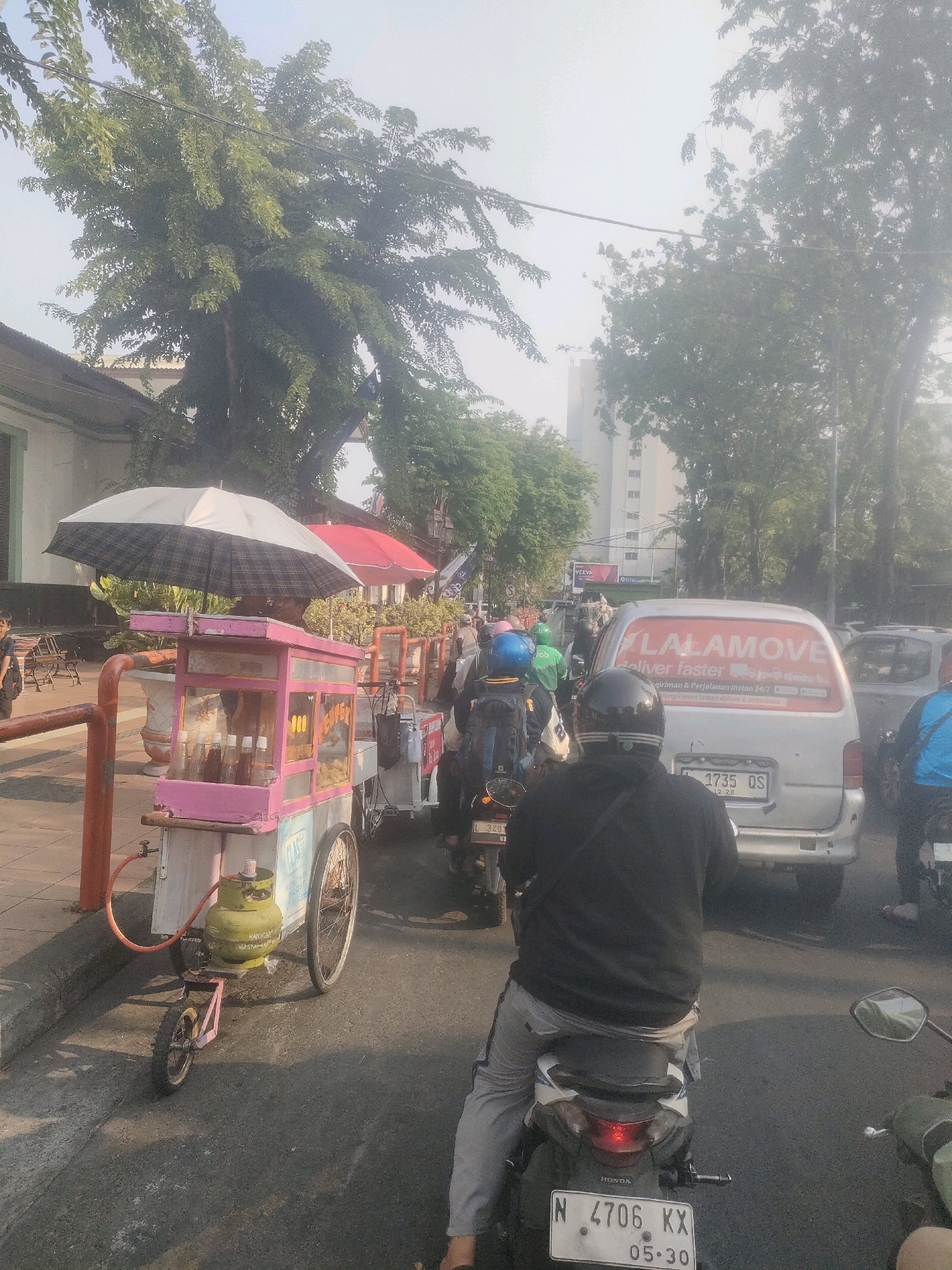 The image depicts a street scene in Surabaya, Indonesia, characterized by a moderate level of traffic. In the foreground, a person is riding a scooter, with their back to the camera. To the left, a pink food cart is parked on the sidewalk, adorned with an umbrella and various bottles. The cart appears to be selling snacks or drinks. Further down the street, a silver van with "LALAMOVE" branding is visible, indicating a delivery service. Several other vehicles and motorcyclists are present, suggesting a busy urban environment. The overall lighting indicates it is daytime, with a slightly hazy atmosphere. The presence of buildings and trees suggests an outdoor, commercial or residential area within the city.