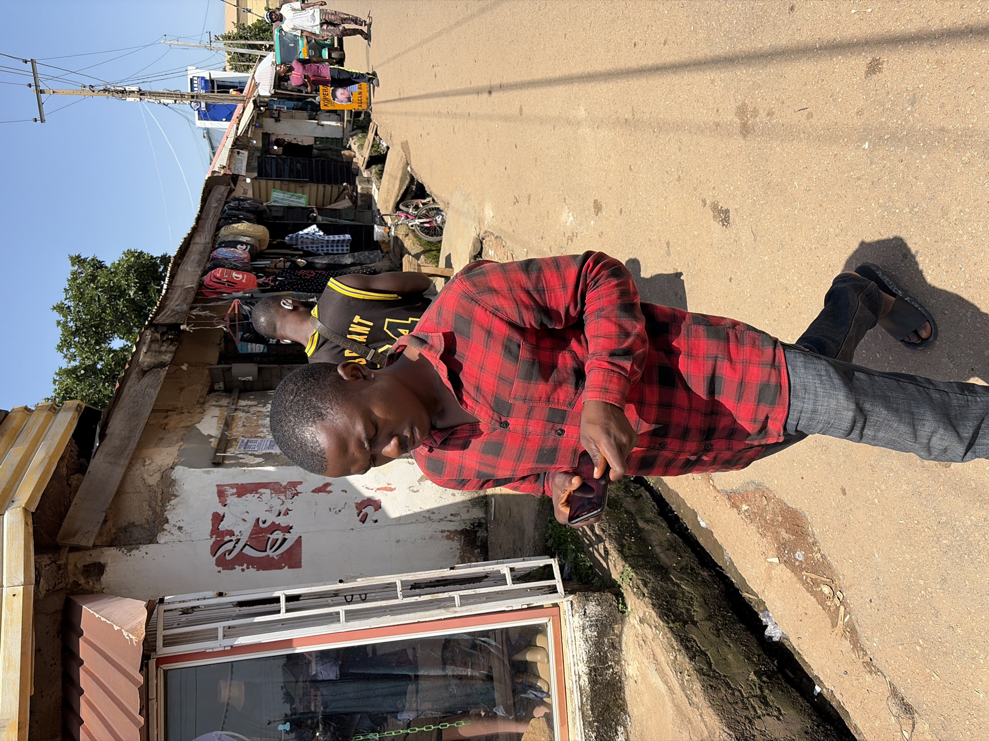 This is a medium shot, eye-level, outdoor photograph taken on a sunny day in Jos, Nigeria. The foreground features a young Black man walking towards the right side of the frame, engrossed in his mobile phone. He is wearing a red and black plaid long-sleeved shirt, dark blue jeans, and black sandals. He is walking on a dirt road that appears to be the edge of a market area.
To the left of the man, there is a small shop with a glass window displaying clothing and mannequins. The shop's exterior is a mix of white plaster and corrugated metal roofing. A red and white sign is partially visible on the wall.
In the background, the scene depicts a bustling market street. Several other people are visible walking and standing. There are more shops with open fronts displaying various goods, including bags and clothing. A bicycle is propped up near one of the shops. Further down the street, a tricycle (keke napep) is parked, and more people are walking away from the camera. Utility poles with numerous wires are prominent in the sky, suggesting the presence of electricity infrastructure. The overall environment appears somewhat weathered and informal, typical of a market area in a developing urban setting. The ground is a mixture of dirt and asphalt, with a drainage ditch running alongside the market stalls.