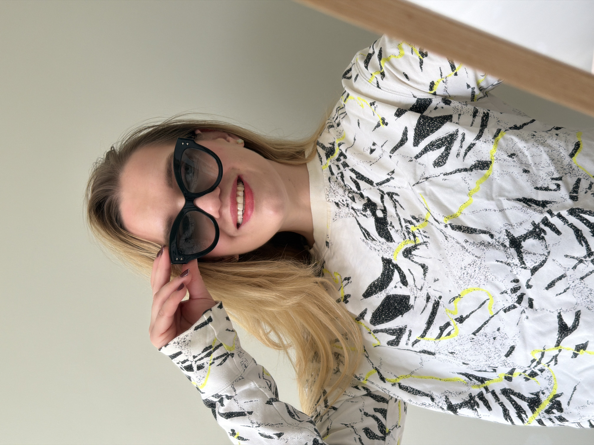 A woman with long blonde hair is wearing black sunglasses and a white blouse with black and yellow patterns. She is smiling. The image is taken at an angle. The background appears to be a plain, light-colored wall. There is no explicit indication that the location is Dreiliņi, Latvia, but that information was part of the request.