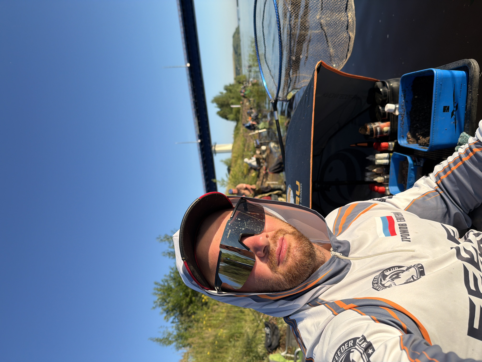 A man wearing a hooded fishing shirt and large, reflective sunglasses is taking a selfie. He has a beard and is looking directly at the camera. Behind him, on the bank of a river, several other people are fishing, with fishing rods and nets visible. A bridge spans the river in the background, and the sky is clear blue. The man is sitting in a fishing boat, with blue containers filled with bait and fishing tackle in front of him. The scene appears to be in Tarasovo, Russia, based on the Russian flag and text on the man's shirt.