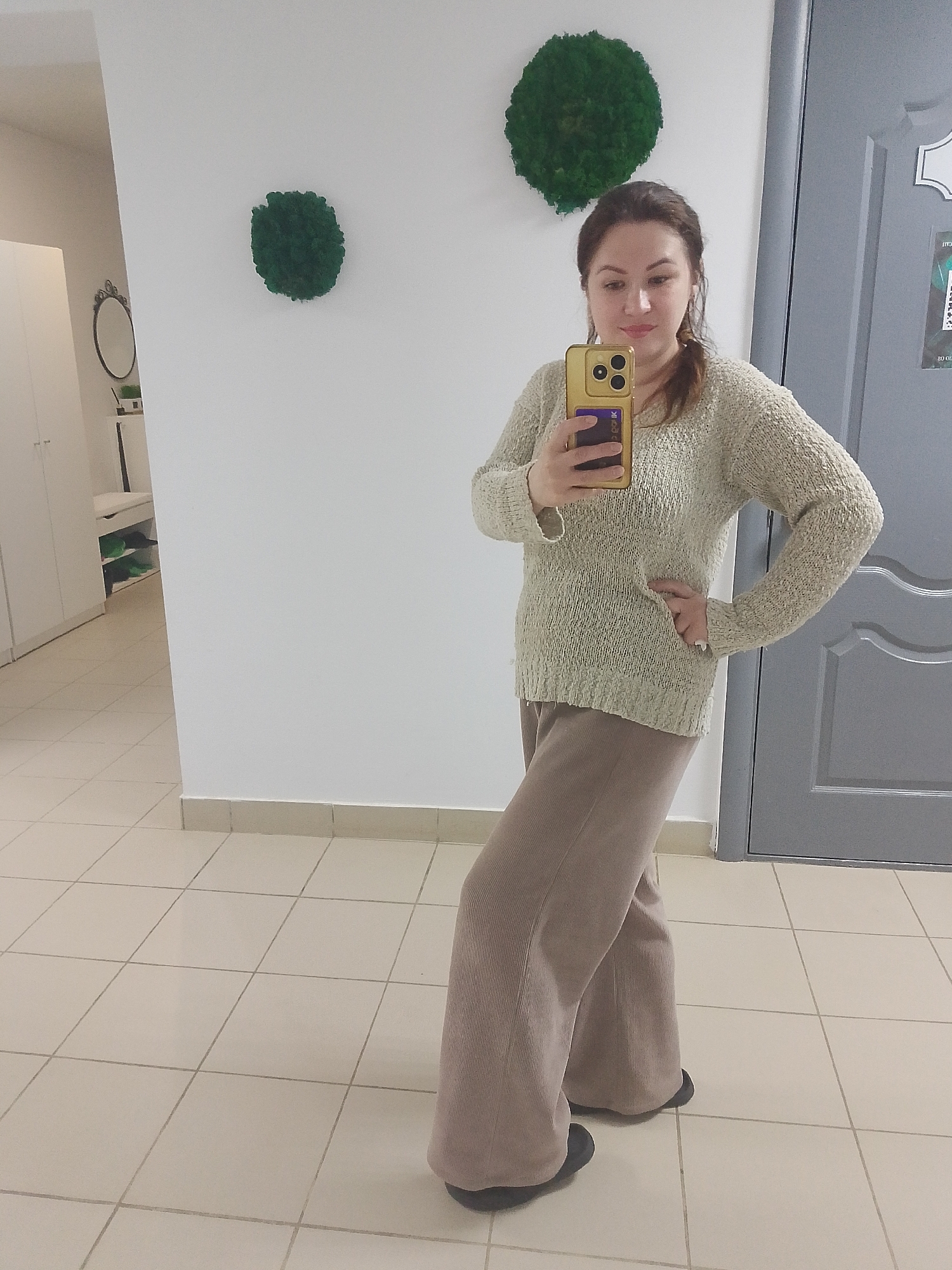 In an indoor full-body mirror selfie taken in Chaikovsky, Russia, a woman stands facing forward, slightly angled to her right, holding a gold-colored smartphone with a purple/blue card visible in its clear case (featuring unclear white text) to capture her reflection. She has brown hair, appears to be smiling subtly, and is wearing a light-colored, textured long-sleeved sweater paired with wide-leg, light brown or taupe pants, with dark shoes peeking out from beneath. Her left hand rests on her hip.
The setting is a well-lit room with white walls and a light-tiled floor featuring dark grout lines. Two circular, green, moss-like decorative art pieces adorn the wall behind her. To the left, an ornate oval mirror reflects a white cabinet area, where more white cabinets and shelving with some green items (possibly shoes or boxes) are visible. To the right, a grey panelled door with a sticker bearing fragmented, unclear text (including "ITS" and "0_0K" or similar characters) is partially visible. The room has a clean, modern, and minimalist aesthetic, possibly a hallway, dressing room, or commercial space. The lighting suggests daytime or bright artificial illumination.