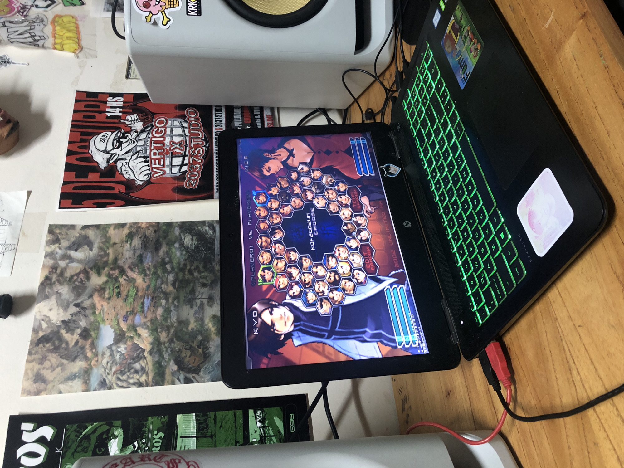 The image shows a laptop on a wooden surface, displaying a fighting game character selection screen, specifically "The King of Fighters 2002 Unlimited Match". The laptop has a green backlit keyboard and stickers on the lid. To the left of the laptop is a white speaker, and various posters and papers are scattered around the area. One poster has the text "VERTIGO 14 HS" and "2037 STUDIO". This image does not contain enough details to determine the location of the city of Buenos Aires, Argentina.