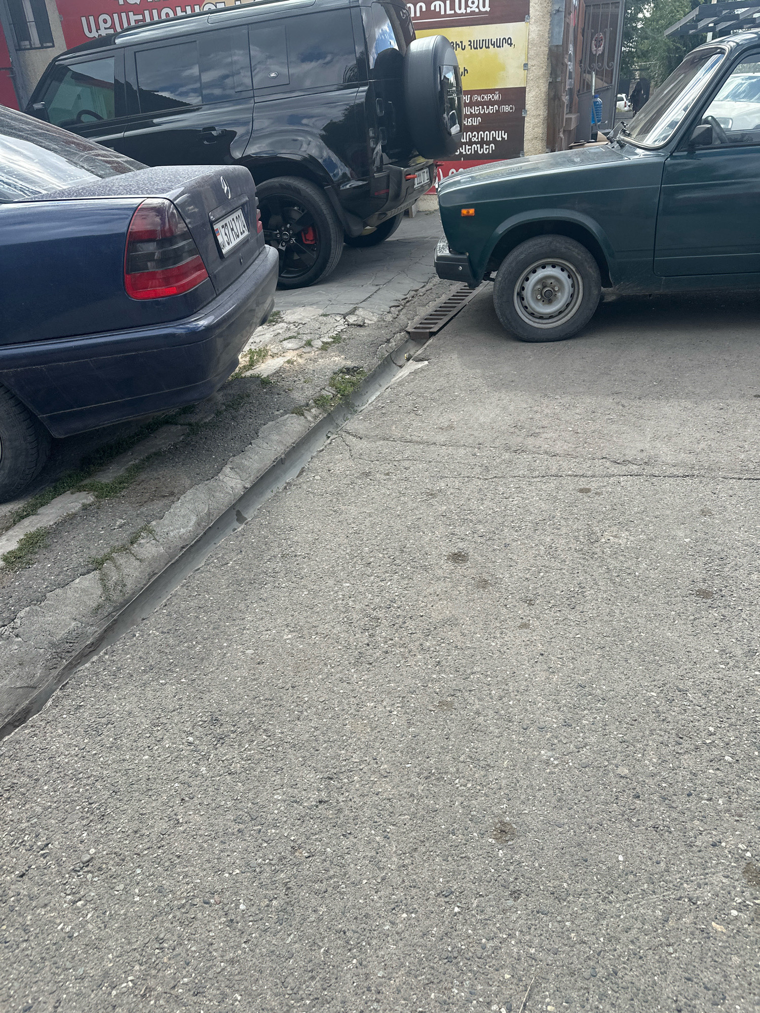 The image shows a street scene in Gyumri, Armenia. In the foreground, a dark blue Mercedes-Benz sedan is parked, with its rear end visible. The car has a license plate reading "37 RH 224". To the right of the sedan, a dark green Lada car is parked. Behind the Mercedes, a black Land Rover Defender SUV is parked, its spare tire mounted on the rear. The cars are parked on an asphalt road with a concrete curb and a drainage ditch. A building with signage in Armenian is visible in the background. The scene appears to be a quiet street, likely in a residential or commercial area of Gyumri. There are no people clearly visible in the image. The lighting suggests it is daytime.