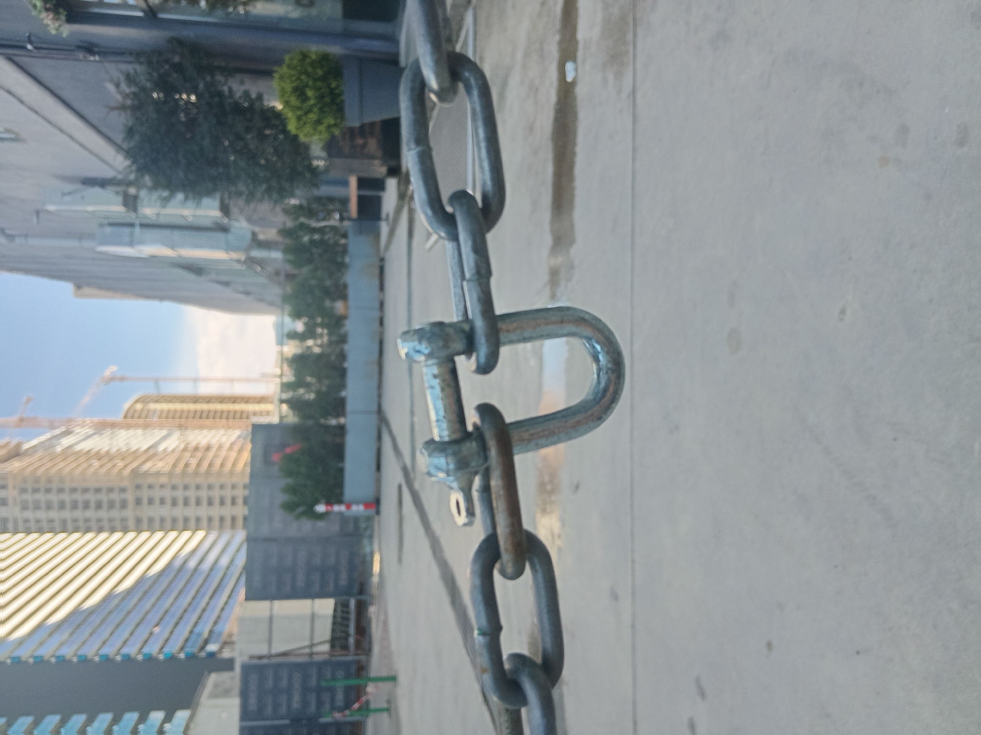 A close-up shot of a metal chain and a U-shaped shackle dominates the foreground. In the background, blurred buildings are visible, including one under construction with cranes. The setting appears to be an urban area, specifically Batumi, Georgia. The ground is a concrete-like surface. There is no one in sight.
