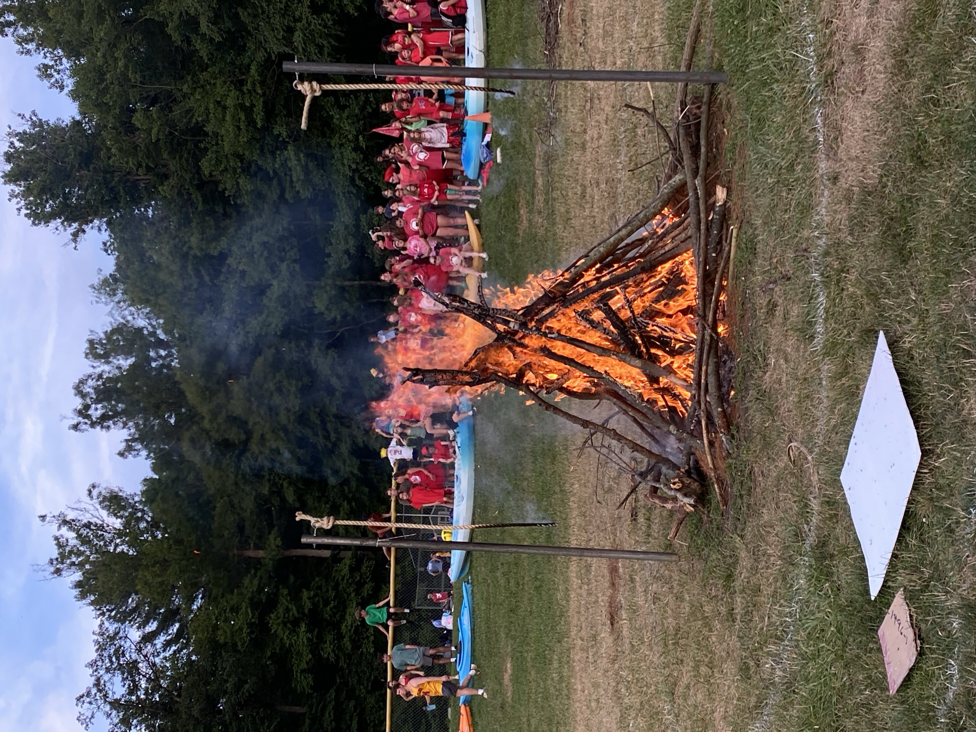 A group of young people, predominantly wearing red, are gathered outdoors near a large bonfire. Many are standing behind a fence or sitting on kayaks. The scene appears to be a recreational event, possibly a camp or a community gathering, set in a grassy area with trees in the background. The sky is overcast with some blue visible. Two metal poles with ropes are positioned in the foreground near the bonfire. The bonfire is actively burning with flames and smoke. In the foreground, there are two flat objects on the grass, one appears to be a piece of cardboard and the other is a white square object. The image does not provide enough information to determine the specific location within Washington Township, United States of America.