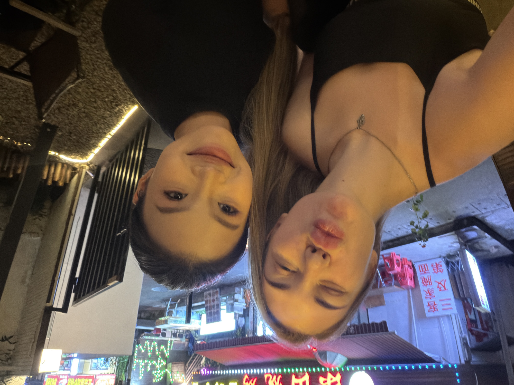 Two young women are taking a selfie in front of a restaurant in Longgang District, China. The woman on the left has long blonde hair and is wearing a black tank top and a silver necklace. She is puckering her lips. The woman on the right has dark hair and is wearing a black t-shirt. She is smiling. The restaurant has a red awning and is lit with neon lights. There are several tables and chairs outside the restaurant. The ground is covered with gravel.