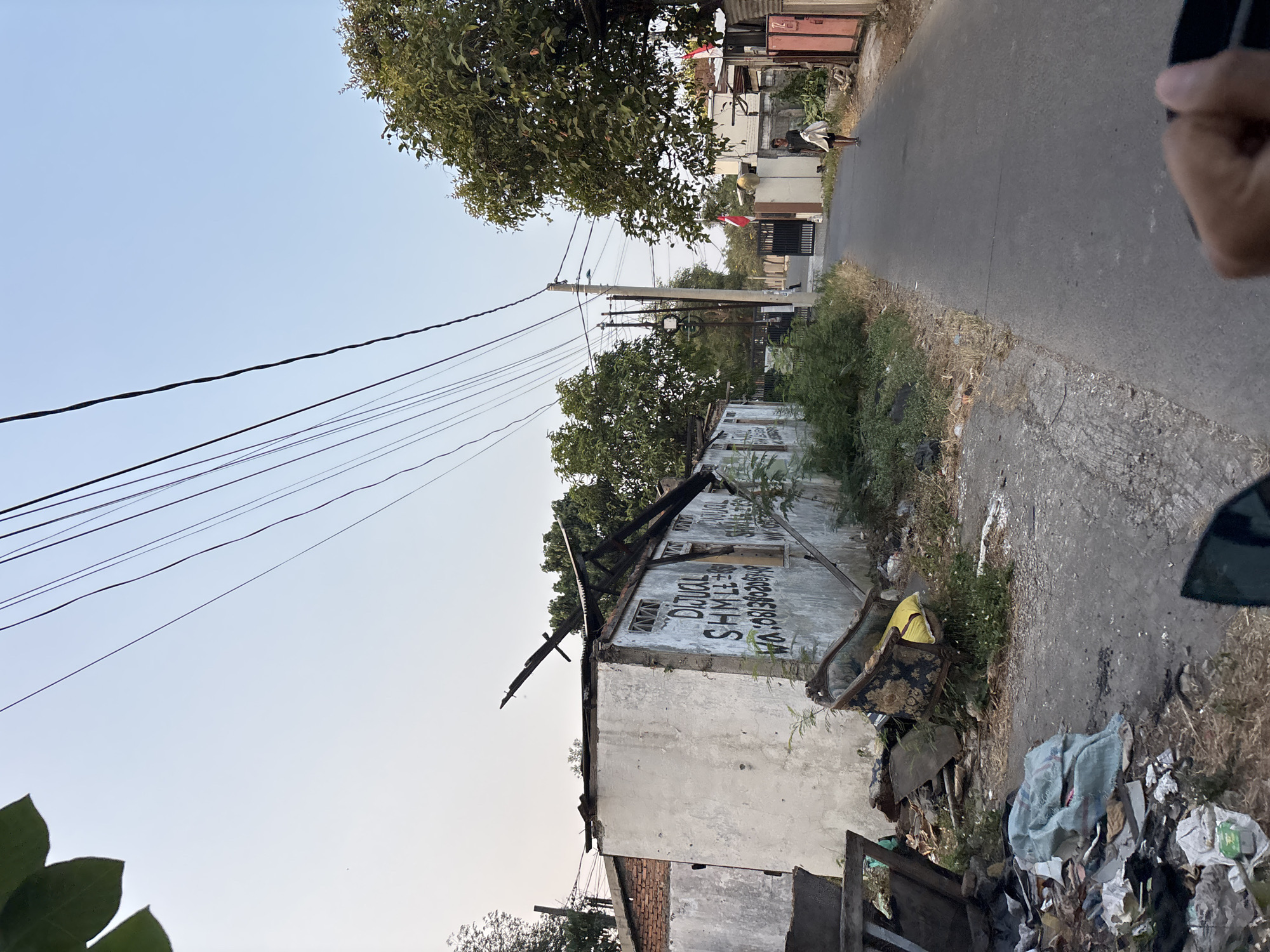 The image depicts a street scene in the city of Bekasi, Indonesia, with a partially damaged building on the left and a residential area on the right. The building on the left, with a severely damaged roof, has the words "DIJUAL" (for sale) and a phone number spray-painted on its exterior wall, suggesting it is being offered for sale. Debris, including a damaged armchair with a yellow cushion and various refuse, is scattered in front of the building. The street itself is paved, with power lines running overhead. On the right side of the street, a person is walking away from the camera, carrying a white cloth or garment. The environment appears to be a typical Indonesian neighborhood with houses and trees lining the street.