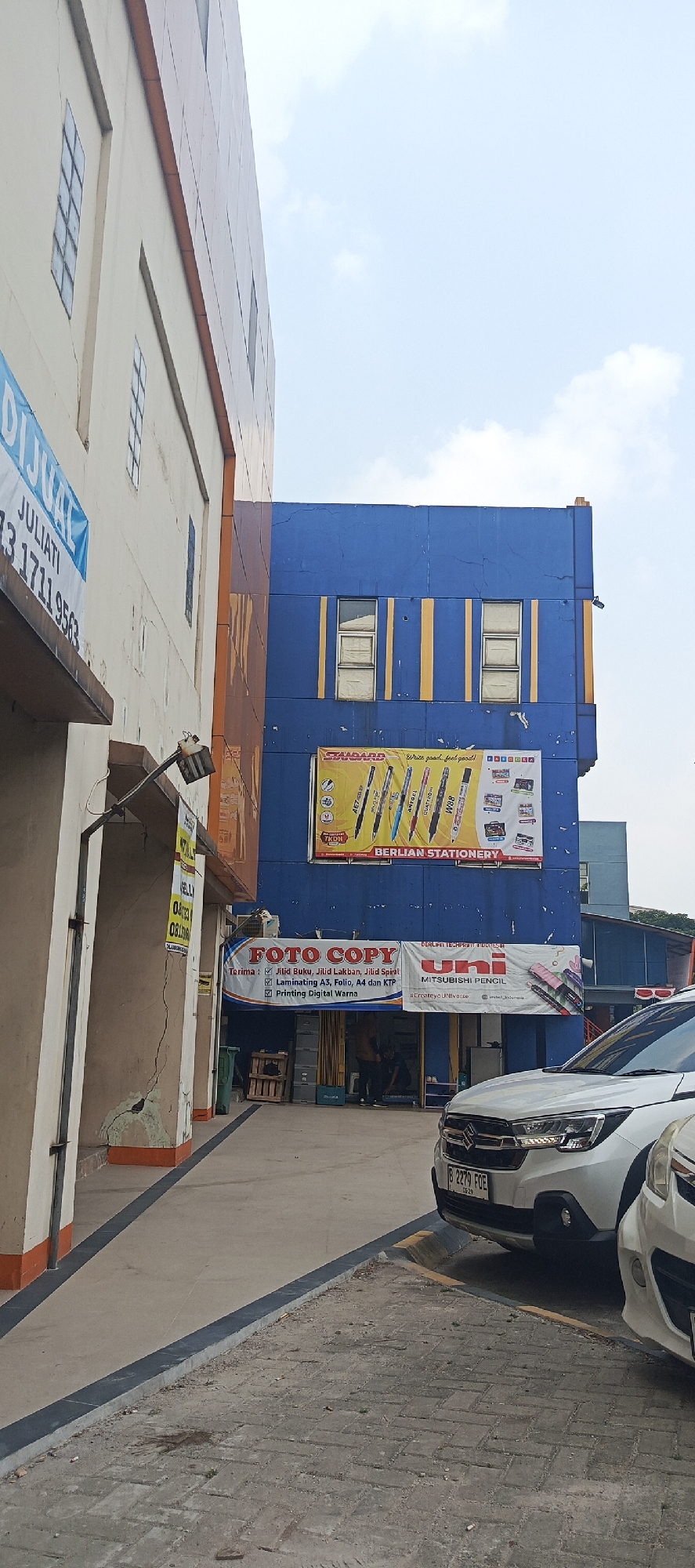 The image shows an exterior view of a building complex in Sukaresmi, Indonesia, under a partly cloudy sky. On the left, a light-colored building with orange trim features several rectangular windows and a blue sign that reads "DIJUAL JULIATI 1711 9563". To the right, a vibrant blue building displays a large banner advertising "BERLIAN STATIONERY" with an image of various pens and a slogan. Below this, another banner announces "FOTO COPY" services, listing "Jilid Buku, Jilid Lakban, Jilid Spiral", "Laminating A3, Folio, A4 dan KTP", and "Printing Digital Warna". A third banner for "UNI MITSUBISHI PENCIL" is also visible.
The ground level of the blue building features a shop entrance with wooden planks inside. Two individuals are partially visible inside the shop, engaged in activity. In the foreground, two white cars are parked. The license plate of the closer car reads "B 2279 FOE". The ground is paved with concrete and interlocking brick sections, with a dark grey border. Green trash bins are present, and some debris is scattered on the pavement. The overall scene depicts a commercial area with businesses catering to stationery and copy services.