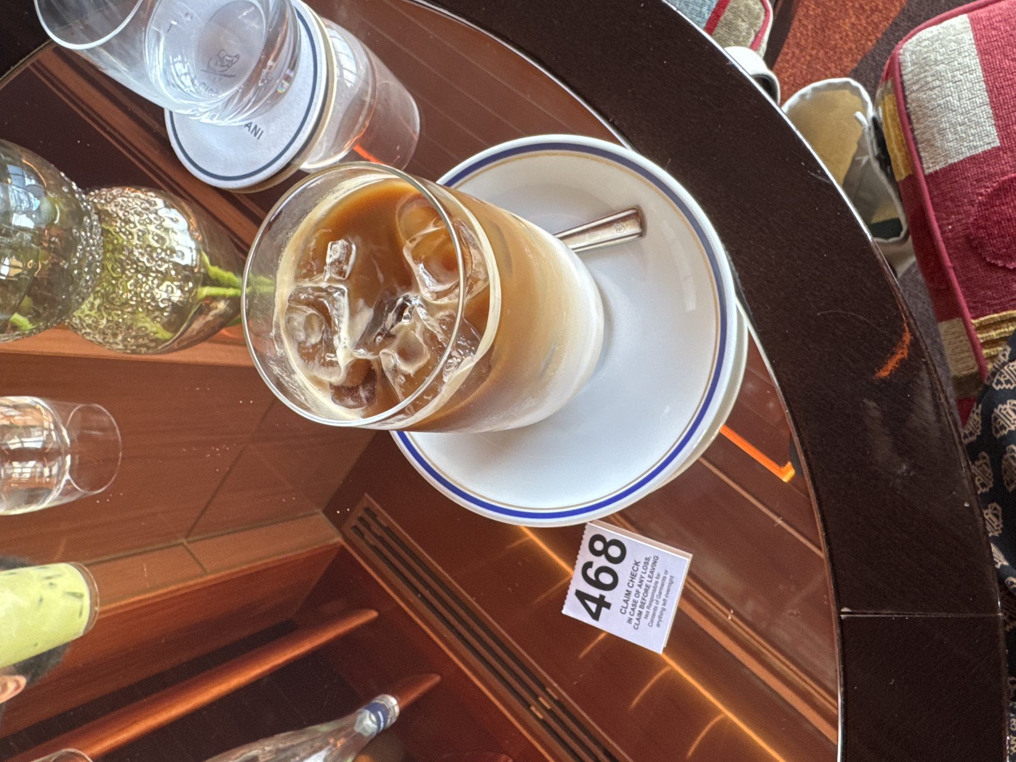 An elevated close-up photograph captures a selection of beverages and objects on a highly reflective, polished dark wood table. The primary subject is a clear glass of iced coffee, featuring distinct layers of milk and coffee with ice cubes, resting on a white saucer with blue and gold rim details. A small silver spoon lies in the saucer. To the left, a white paper claim check ticket, numbered "468," is visible, stating "CLAIM CHECK IN CASE OF ANY LOSS, CLAIM BEFORE LEAVING Not Responsible for Contents of Garments or anything left overnight." Other items on the table include a tall glass with a green iced beverage, an inverted clear glass, another clear glass on a white coaster partially displaying "ANI" text, and a textured, reflective decorative glass object. Reflections of vertical architectural elements are apparent on the table surface. The immediate foreground and lower background feature blurred elements of a patterned, upholstered seating area in shades of red, beige, and dark brown. The sophisticated setting, particularly the "ANI" branded coaster, indicates an establishment associated with the Armani brand, likely Armani/Ristorante at 717 Fifth Avenue in New York, United States. No individuals are visible.