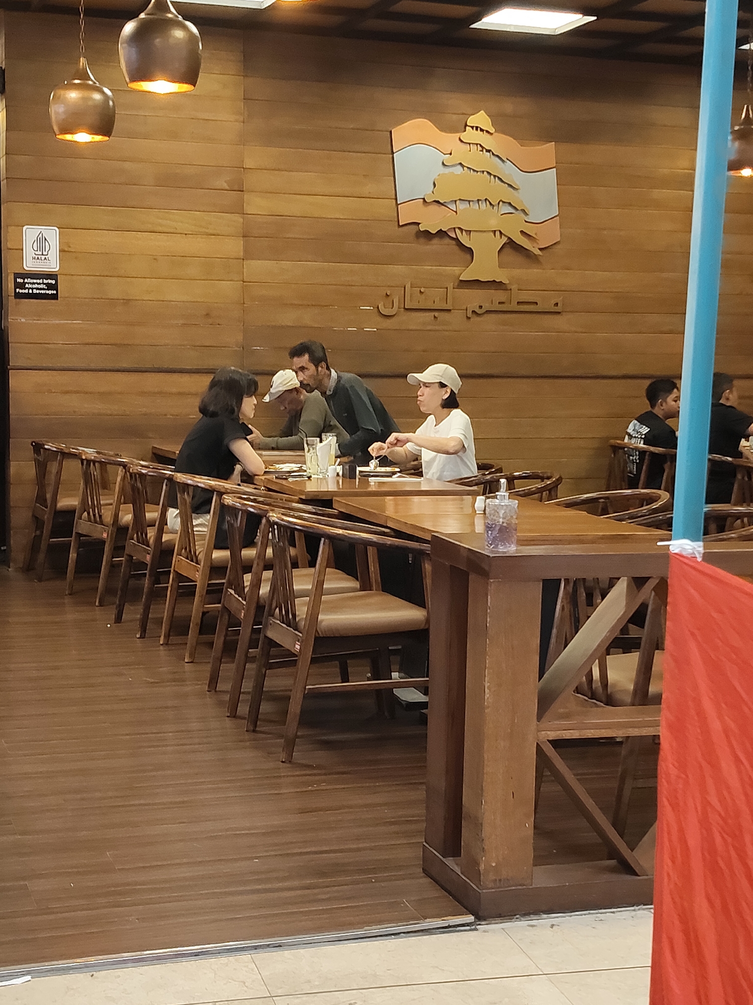 In a restaurant with wooden walls and floor, two women and two men are seated at tables. The woman on the left is wearing a black shirt, and the woman on the right is wearing a white cap and t-shirt. A man in a gray shirt is seated with the woman in black, while another man in a black shirt is seated at a table in the background. Hanging lamps illuminate the dining area. A sign on the wall indicates that the establishment is Halal and does not permit outside food or beverages. The restaurant is located in the city of Medan, Indonesia.