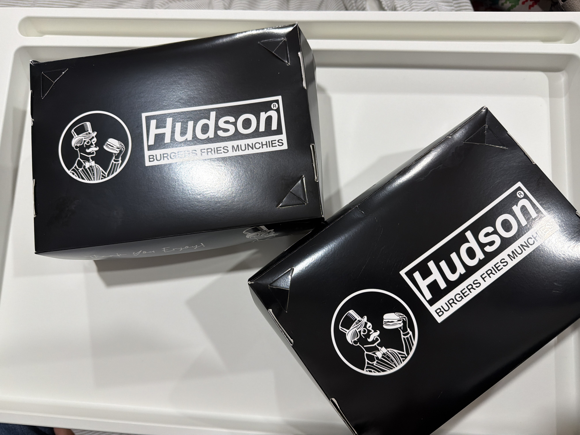 Two black Hudson Burgers Fries Munchies boxes are placed on a white tray. The Hudson logo is displayed on each box in white lettering, along with a circular emblem of a man in a top hat holding a burger. The boxes appear to be made of cardboard and have folded corners. The tray is smooth and clean. The scene is well-lit.