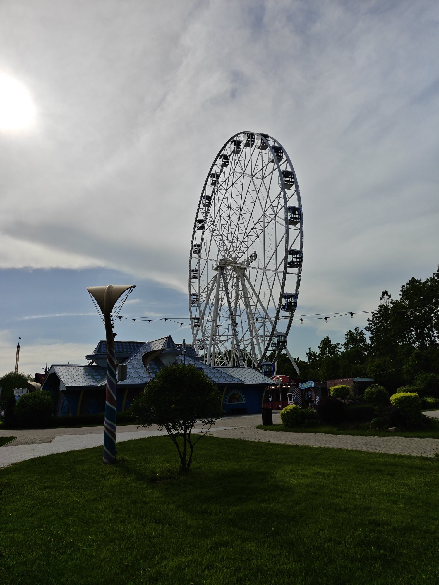 The image depicts a Ferris wheel in a park setting. The Ferris wheel is tall and white with passenger cars. It is located behind a building with a blue and gray roof. A decorative lamp post is on the left of the frame. Lush green grass is in the foreground. The sky is cloudy with the sun visible in the upper left corner. The scene is set in Khimki, Russia.