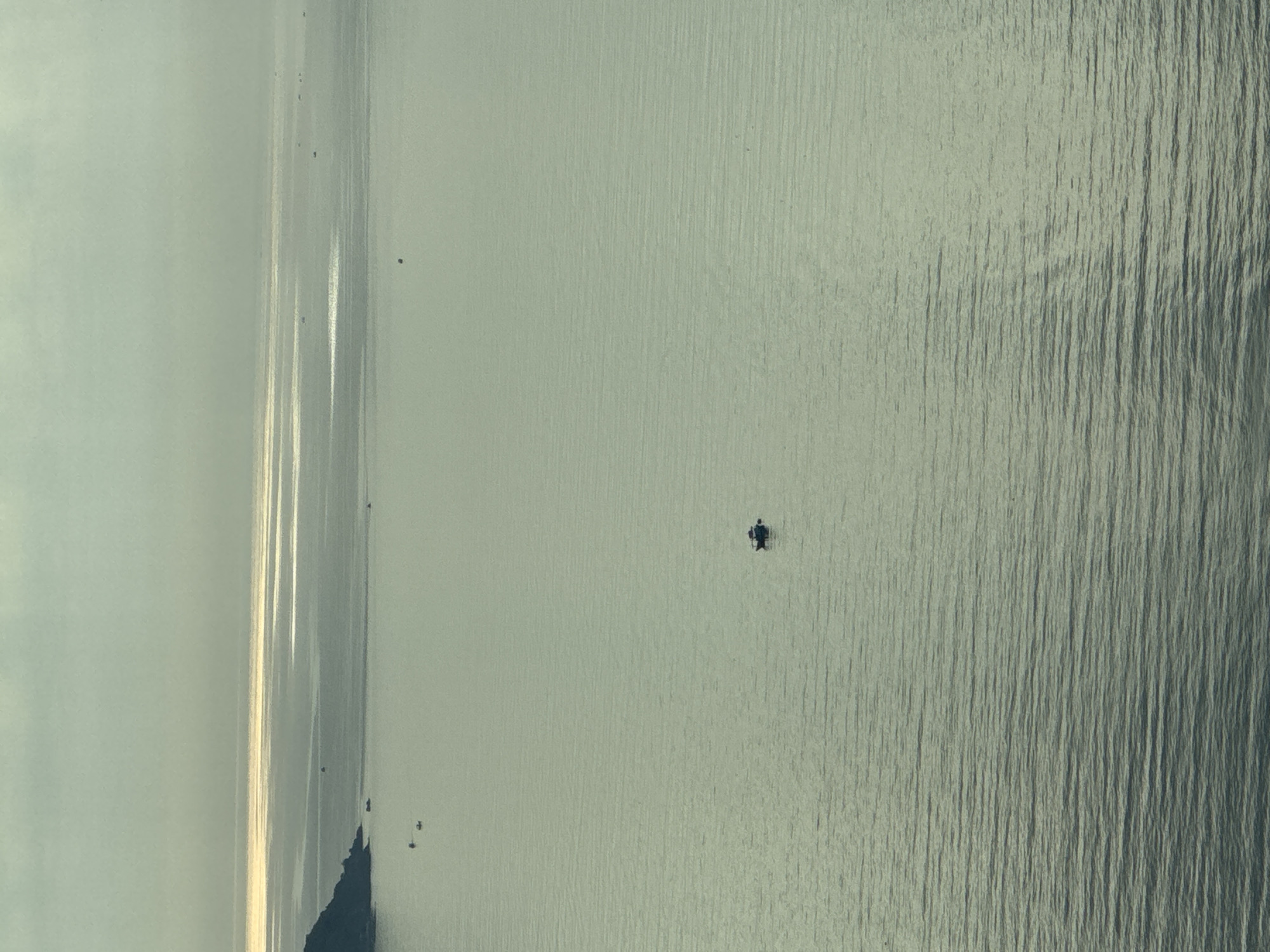The image depicts a body of water, likely the sea, in Bac Nha Trang Ward, Vietnam. A dark figure, potentially a person on a small watercraft, is located in the central portion of the water. The left side of the image shows the coastline, with faint reflections of sunlight shimmering on the water's surface. The overall tone is muted and grey.