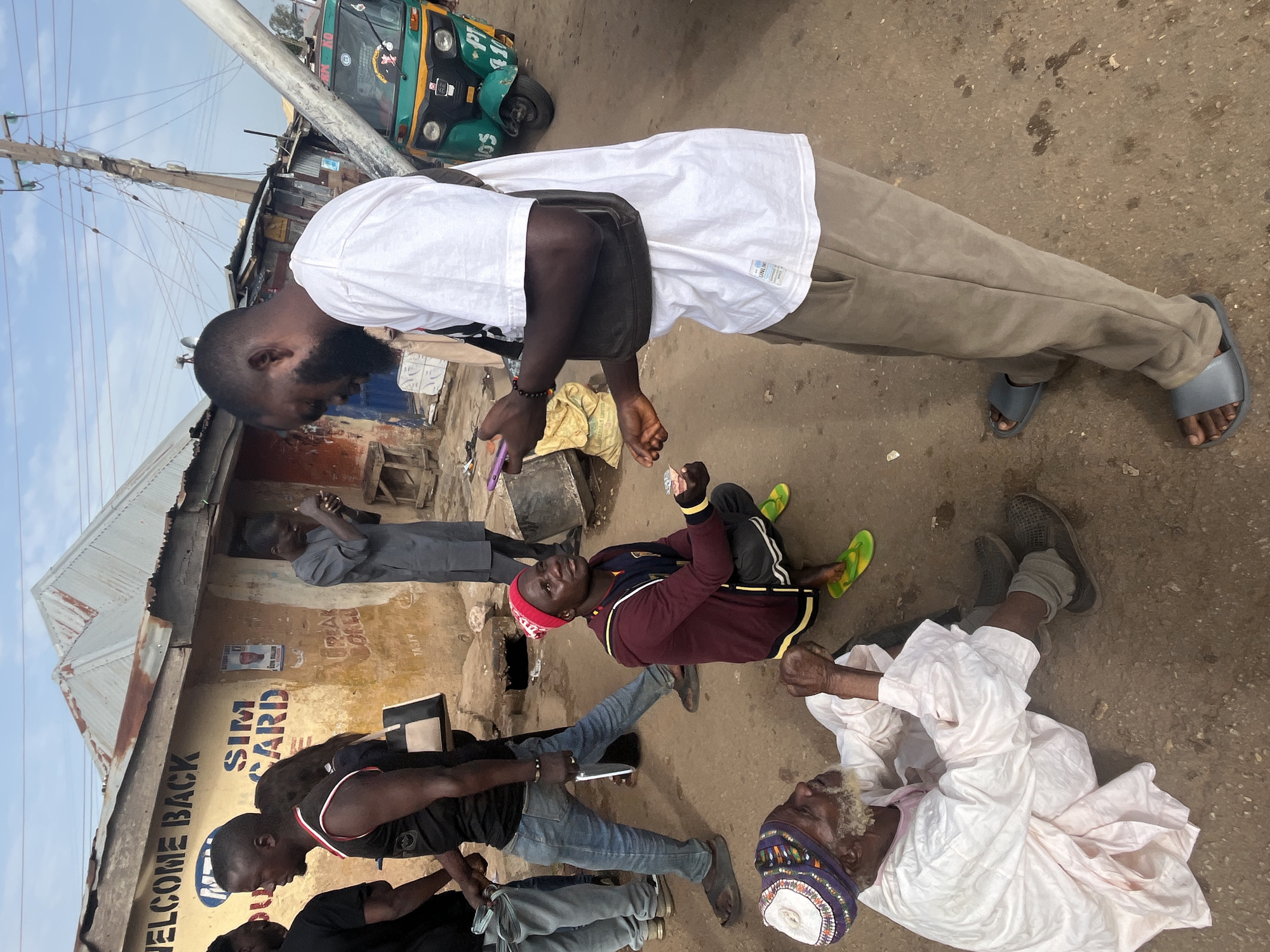 In Gura Top, Nigeria, an outdoor scene unfolds with several individuals present. In the foreground, an elderly man with a white beard, wearing a light-colored robe and a patterned cap, is seated on the ground. To his right, a younger man in a red beanie, maroon jacket, and dark pants is kneeling. He appears to be engaged in a transaction, holding out money to the man standing over him. This standing individual, dressed in a white t-shirt and tan pants, is looking down at his phone and extending his hand, seemingly receiving the money. A second younger man, wearing a black tank top and blue jeans, stands behind the kneeling man, holding a wad of cash. Further in the background, other individuals are visible, including one in a grey traditional garment standing near a building with signage. A brightly colored motorized tricycle is parked on the right side of the frame. The environment suggests a dusty, urban street with a simple building featuring corrugated metal roofing and various advertisements on its wall.