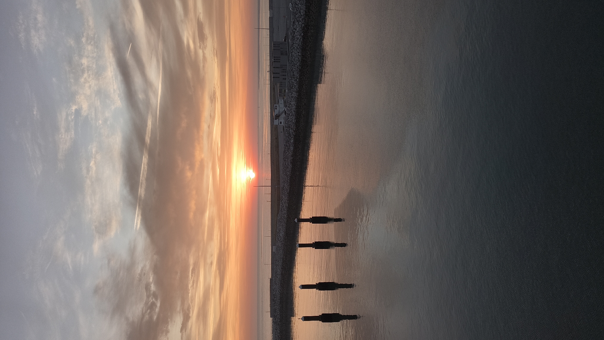 The image captures a serene sunrise over the port of Calais, France. The sun is positioned low on the horizon, casting a warm orange glow across the sky and reflecting on the still waters of the harbor. Wispy clouds, tinged with hues of pink and orange, stretch across the sky. In the foreground, four dark posts rise from the water, their reflections visible below. Beyond them, a stone pier runs along the edge of the harbor, leading to buildings and infrastructure typical of a port. The overall scene is tranquil and picturesque, highlighting the natural beauty of the sunrise in an industrial setting.