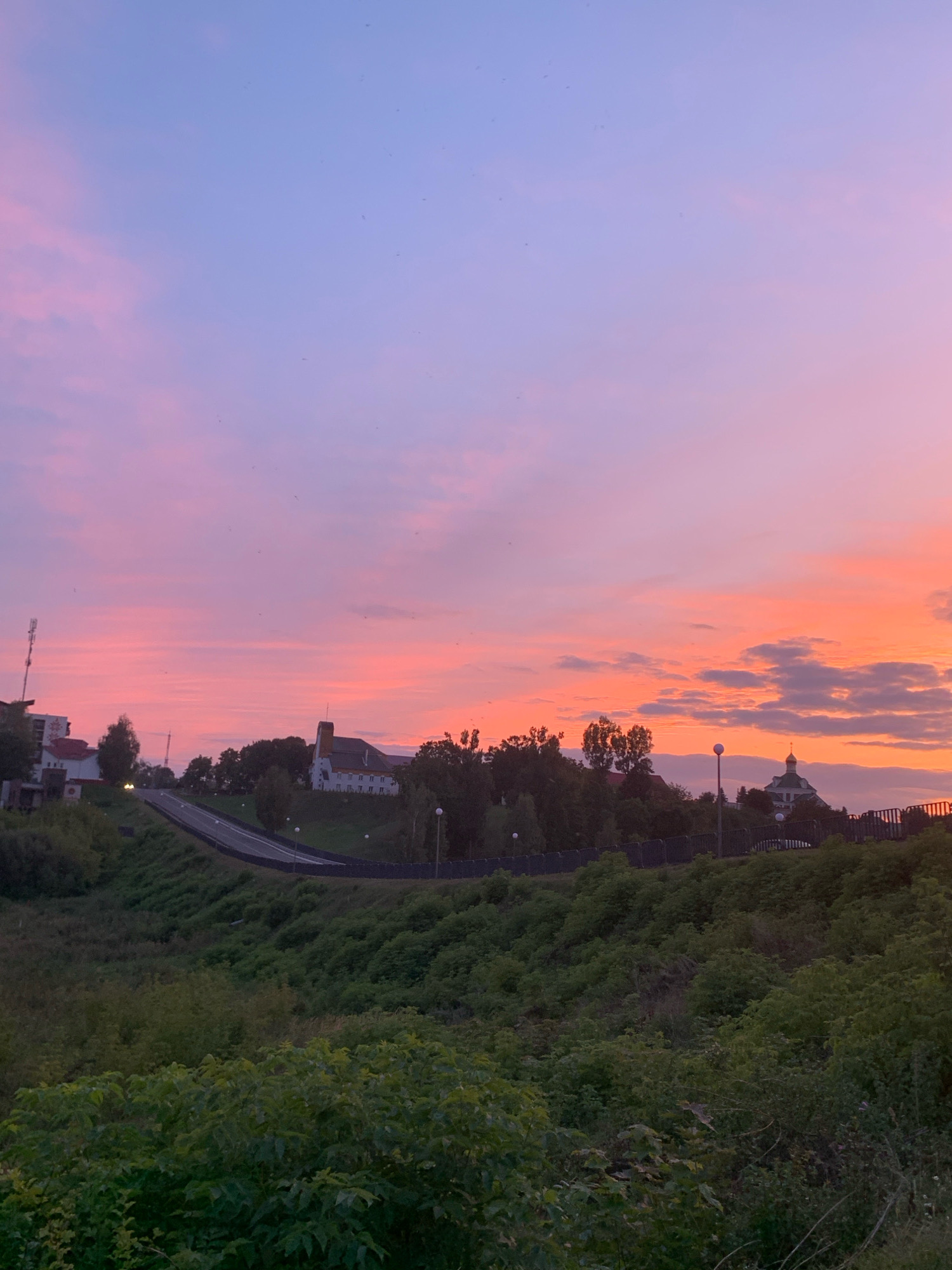 The image depicts a sunset scene in the city of Kapyĺ, Belarus. The sky is a gradient of soft purples and pinks, with wispy clouds. In the mid-ground, a paved road winds its way up a grassy hill. Several buildings are visible along the hillside, including a white building with a red roof, another larger white building with a tower, and a domed structure. Trees and lush green foliage occupy the lower portion of the image. The overall impression is one of a tranquil, rural landscape at dusk.