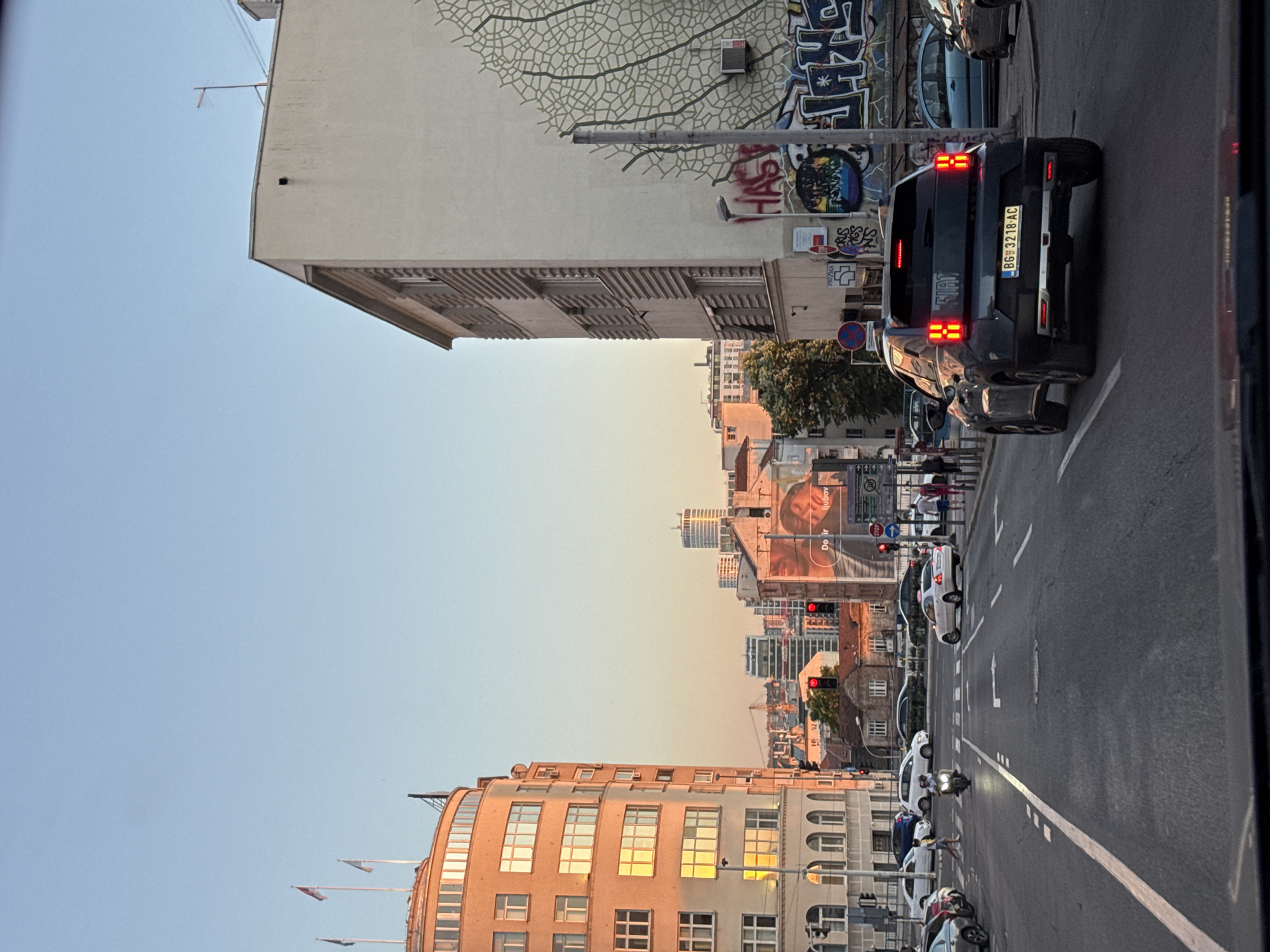 The image captures a street scene in Belgrade, Serbia. The view looks down a street lined with buildings of varying architectural styles, ranging from older, ornate structures to modern high-rises in the distance. Graffiti adorns the side of one building, adding an urban element to the scene. Traffic lights are visible, indicating intersections and controlled pedestrian crossings.
A dark Fiat vehicle is parked on the right side of the frame, bearing a license plate "BG-3218-AC", likely registered in Belgrade. Cars are parked along the street. Several people are walking on the sidewalks, some crossing the street. The lighting suggests either early morning or late afternoon, casting a warm glow on the buildings. Flags are flying from the tops of some buildings. The street is marked with lane dividers.