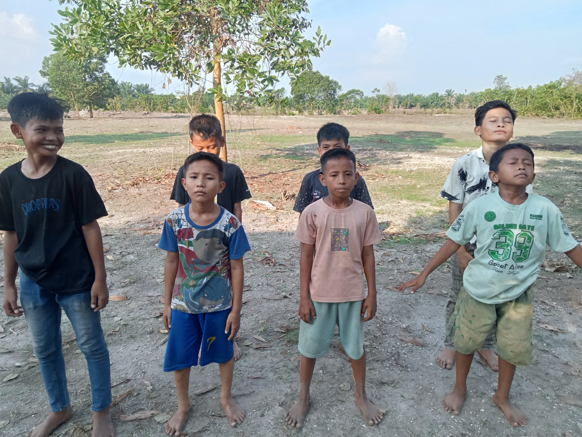 Six boys, appearing to be of Indonesian ethnicity and between the ages of approximately 6 and 13 years old, are standing in a line in an outdoor, rural setting. The ground is dry and dusty, with scattered leaves and debris. In the background, there is sparse vegetation, including trees and palm trees, and a hazy sky with some clouds. The overall scene suggests a location in Teluk Kenidai, Indonesia, characterized by its rural landscape.
From left to right, the first boy is wearing a black t-shirt and blue jeans. He is smiling and looking to his left. The second boy is wearing a black t-shirt and is looking down. The third boy, in the front center, is wearing a blue and white t-shirt with cartoon characters and blue shorts. He is looking forward with a neutral expression. The fourth boy, behind the third, is wearing a dark t-shirt with a pattern and is looking down. The fifth boy is wearing a light pink t-shirt and light green shorts, with his eyes closed and his head tilted back slightly. The sixth boy, on the far right, is wearing a light green t-shirt with graphics and green shorts. He also has his eyes closed and his head tilted back, with his left arm extended to the side. The boys are all barefoot. The lighting suggests it is daytime, possibly late afternoon due to the angle of the sun.